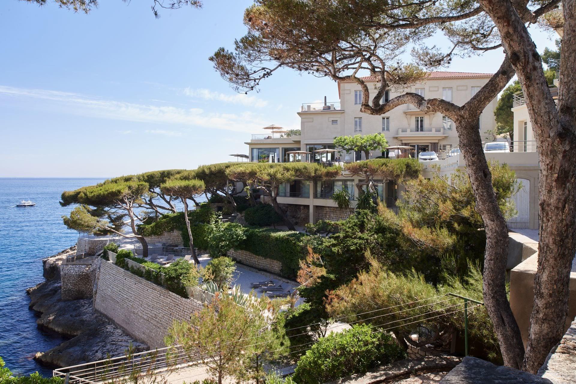 Les Roches Blanches: the hotel building on a rocky coastline overlooking the Mediterranean with a pine tree in foreground