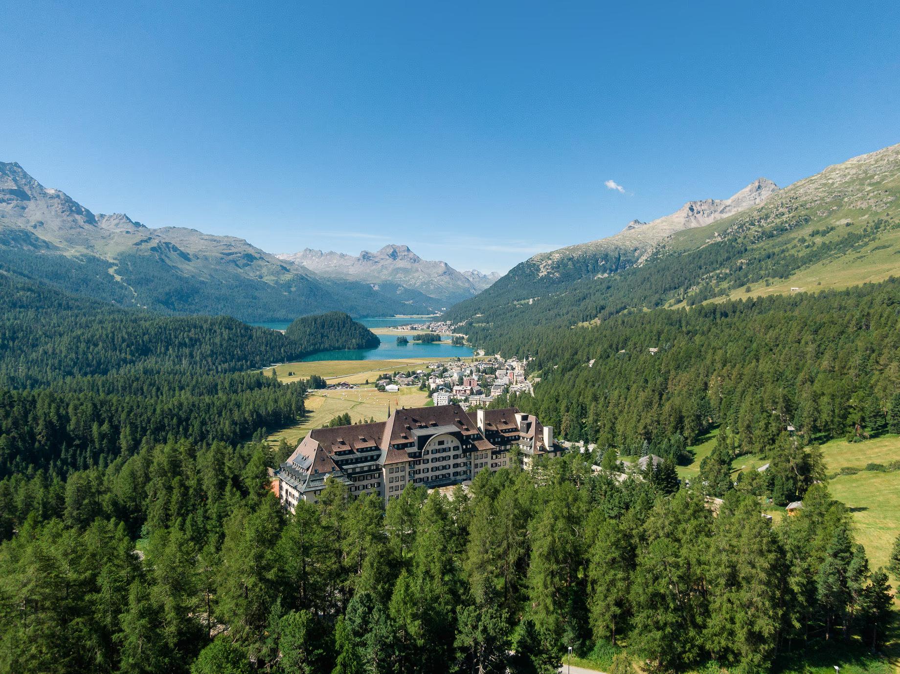 A large hotel in a forest, with a village and a lake in the valley and mountains beyond