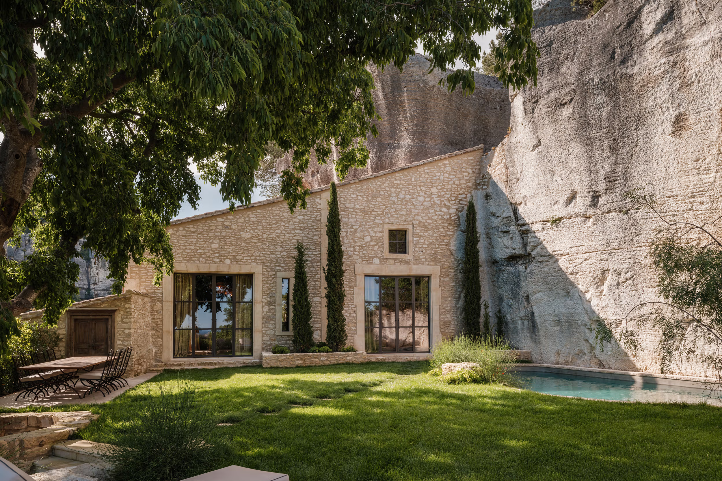 Pale stone villa facade with modern glass doors, cypress plantings, turquoise pool, limestone cliff wall