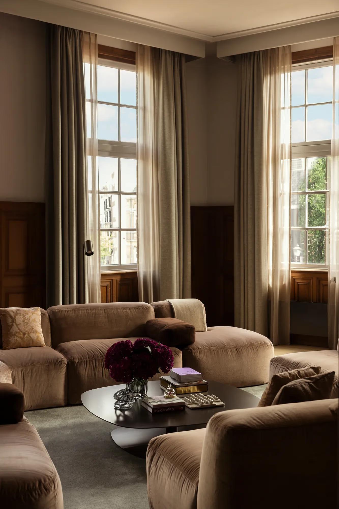 A sitting room with tan velvet sofas, dark red flowers on a low table and tall curtained windows