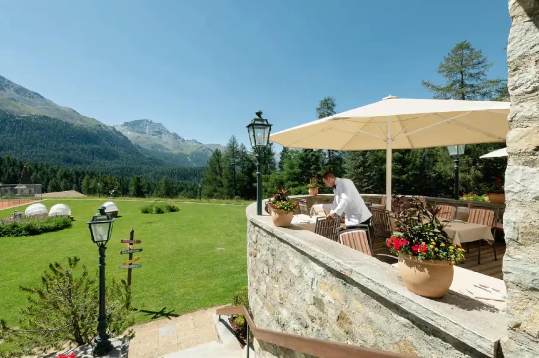 Suvretta House: an outdoor terrace dining area with cream umbrella, verdant lawn, and mountain backdrop