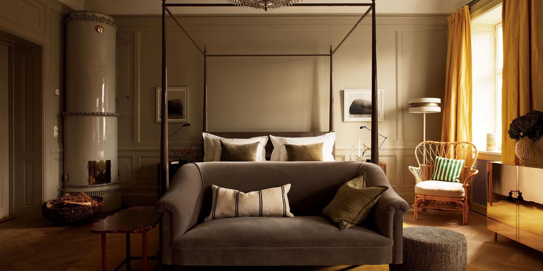 A canopy bed with dark frame and gold cushions in a room with warm neutral walls and tall arched windows