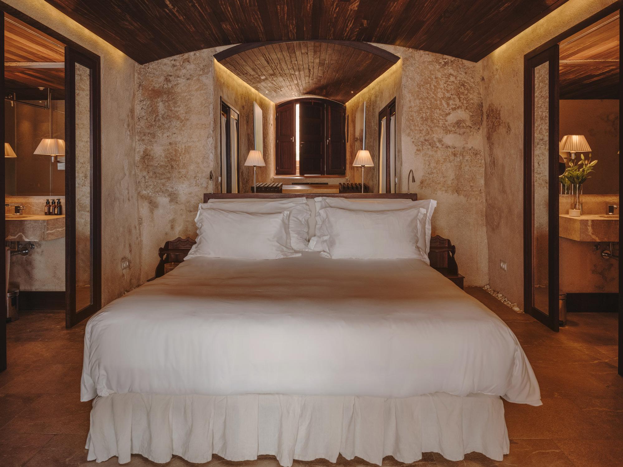 Cap Rocat: a spacious bedroom with a white bed, wooden headboard, pale ochre walls, and wooden ceiling beams