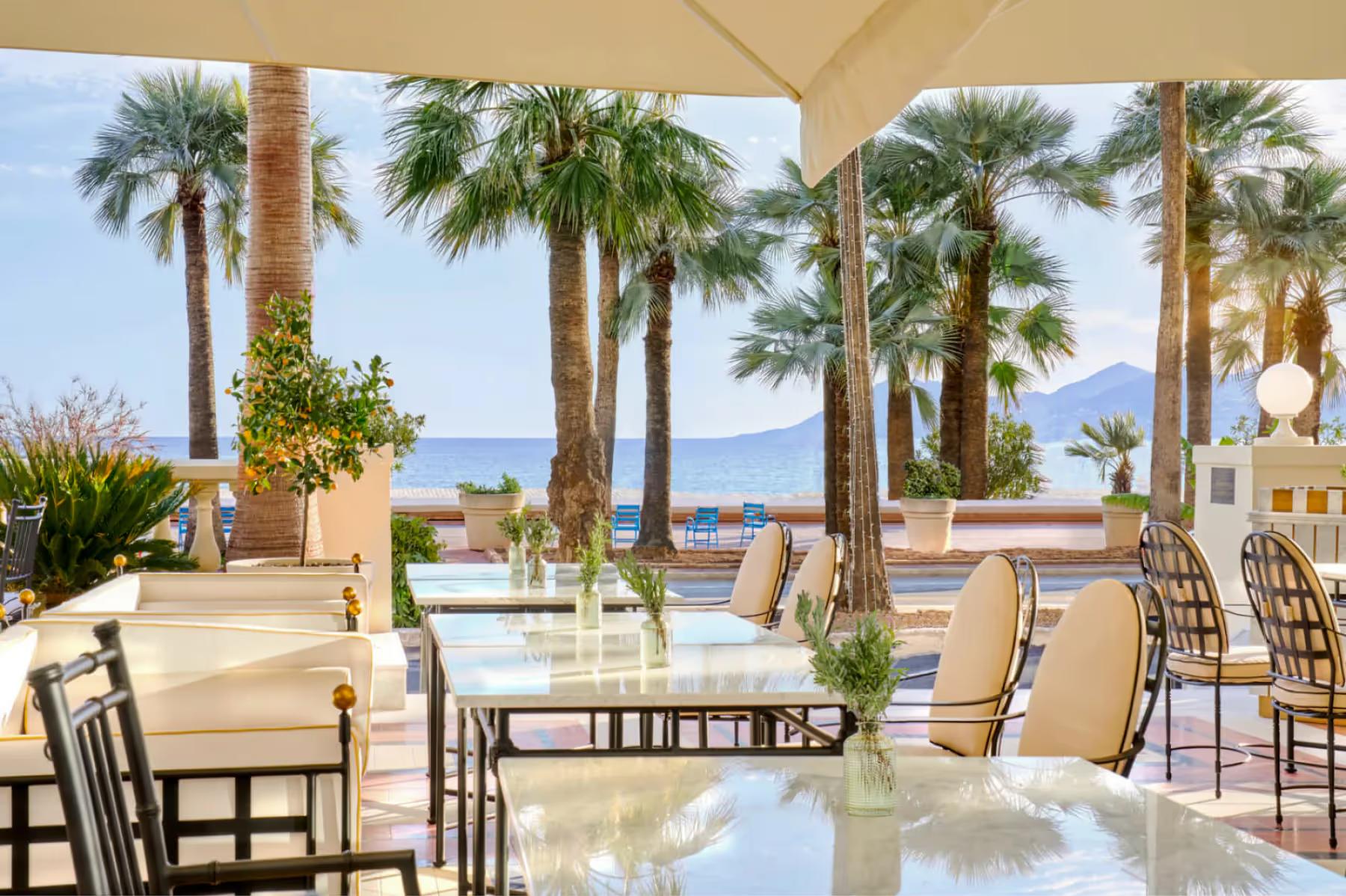 Carlton Cannes: beachside terrace dining with cream-colored umbrellas, palm trees, and sea views beyond