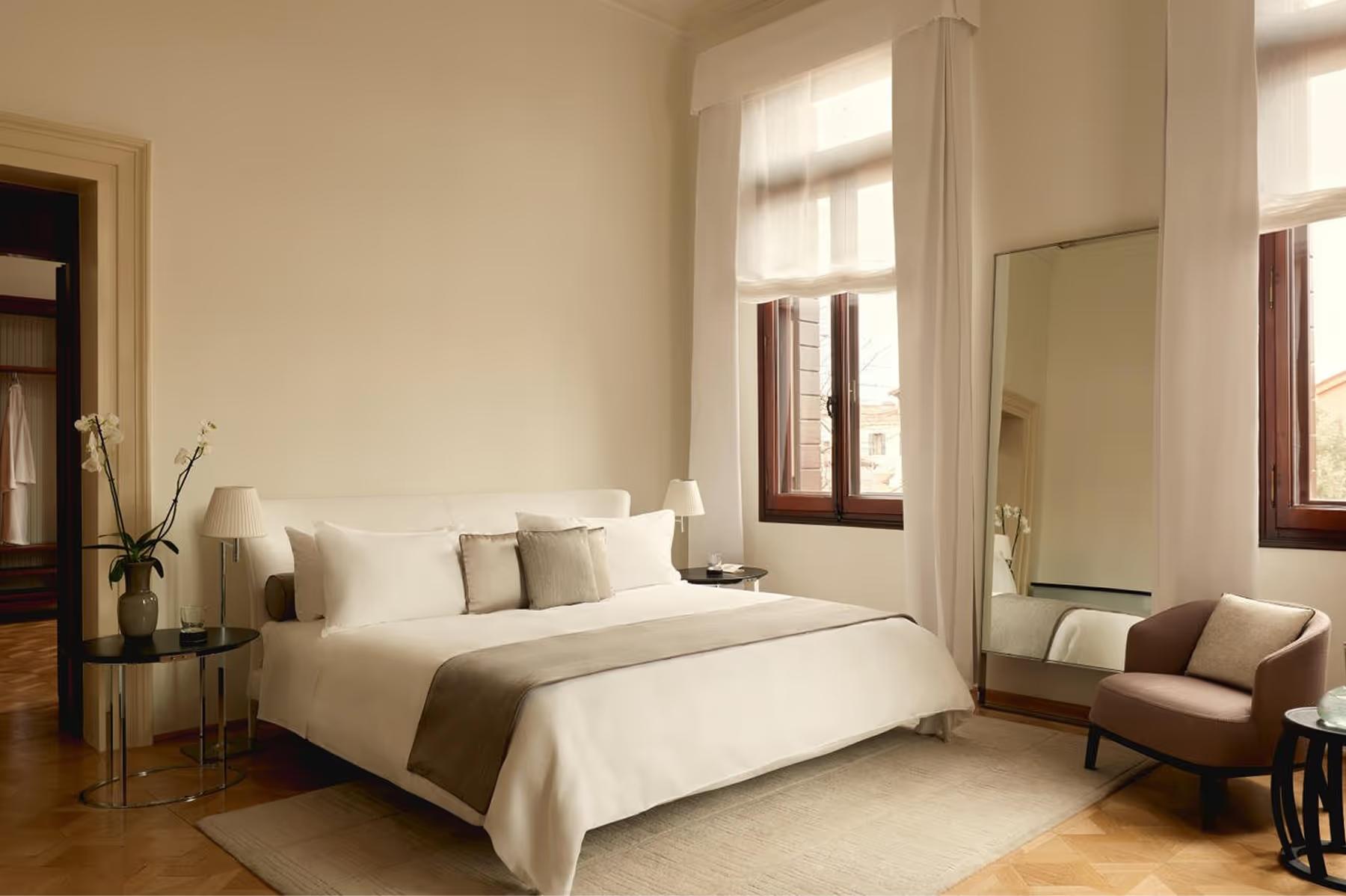Aman Venice: a bright bedroom with neutral linens, brown leather seating, tall windows with natural light, and wooden flooring