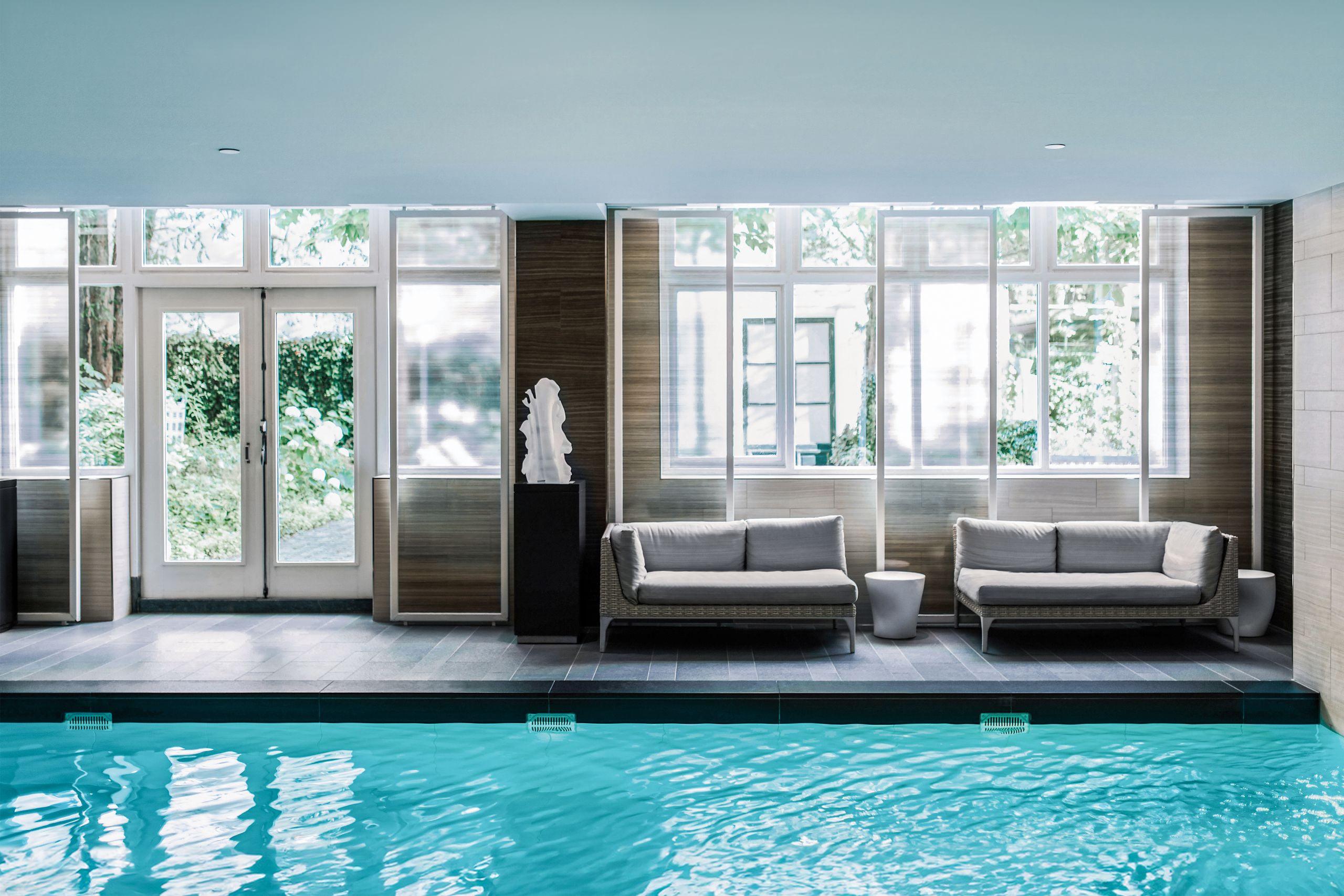 An indoor pool with bright turquoise water flanked by lounge seating and tall windows framed in dark wood