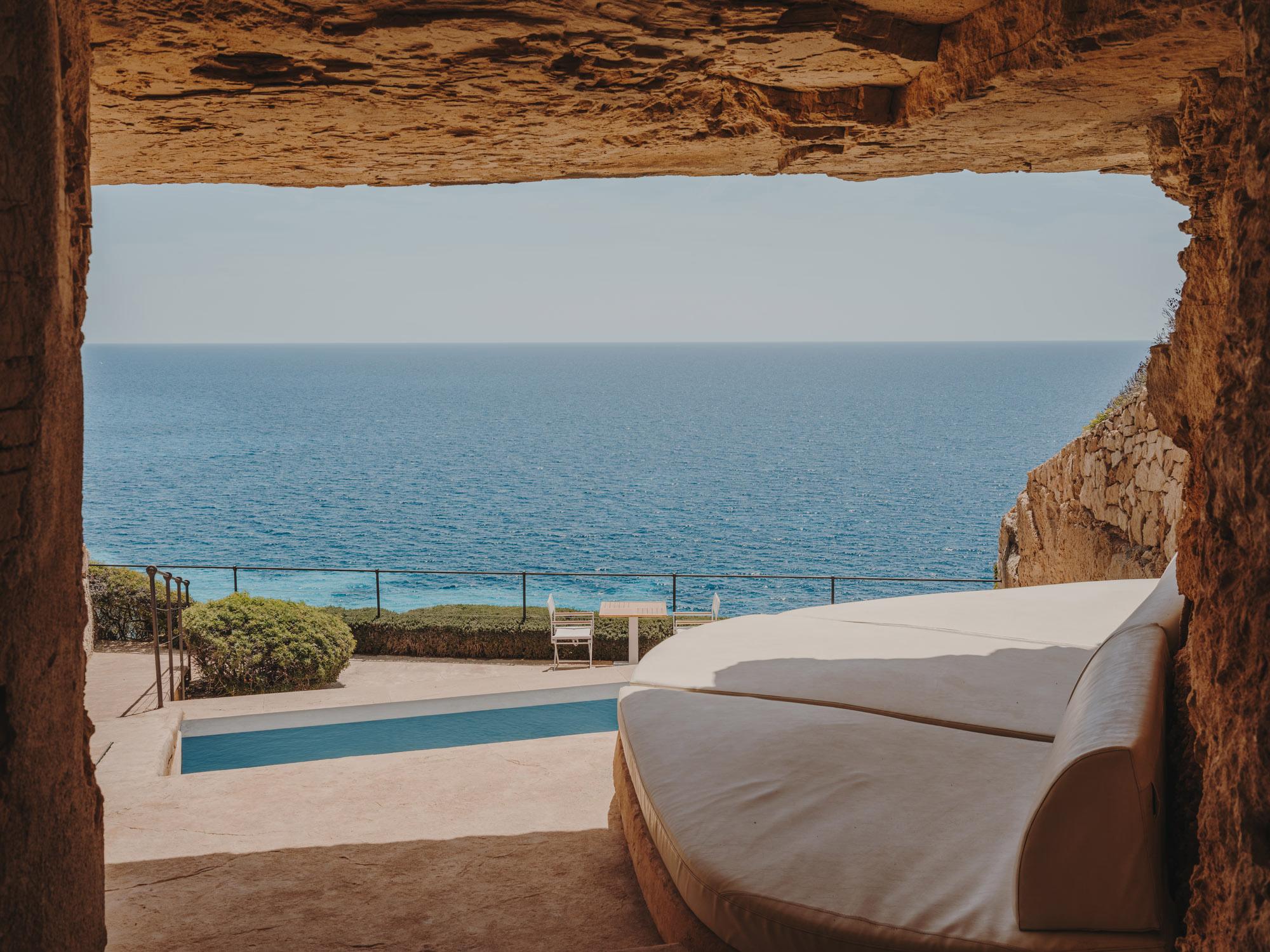 Cap Rocat: a figure in a tan linen coat walks beside a turquoise pool under sun and coastal cliffs
