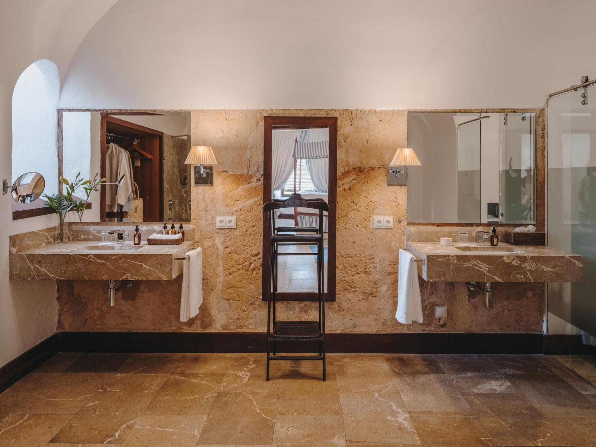 A dual vanity bathroom in warm stone with brass fixtures, mirrors, white towels, and pale archways