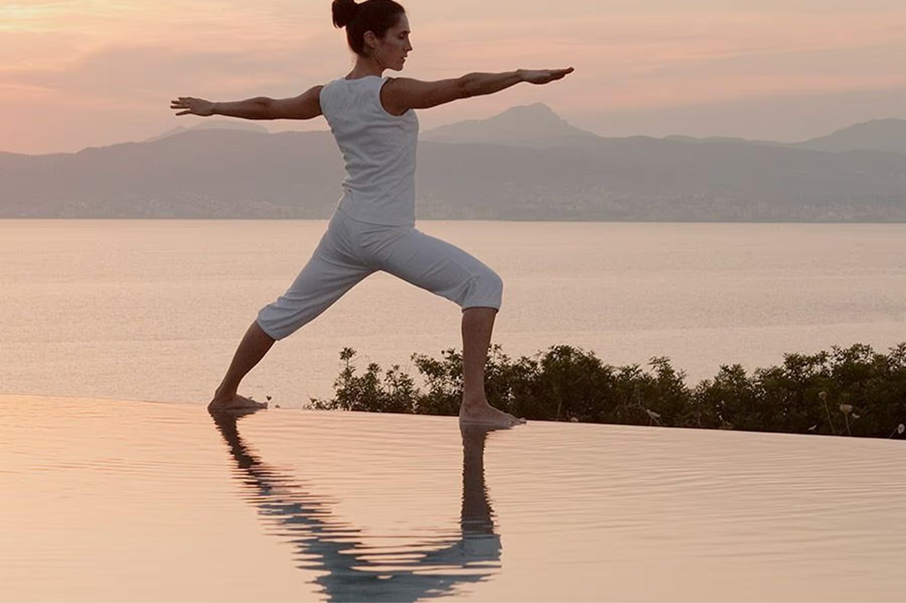 A figure practices yoga in a wide stance pose beside a reflecting pool at golden hour