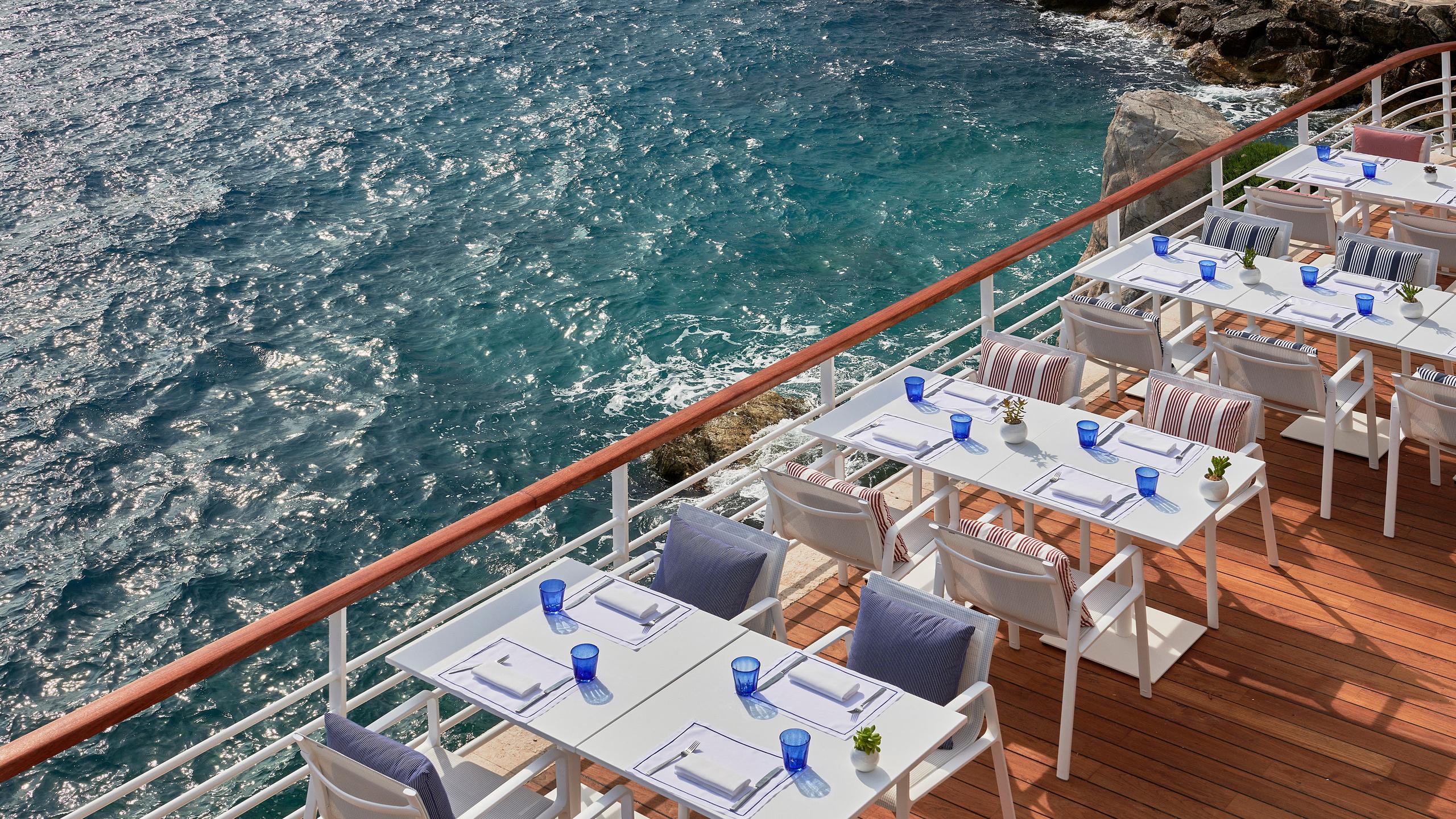 A seaside dining platform with white table settings, water views, and sailboats in the bay
