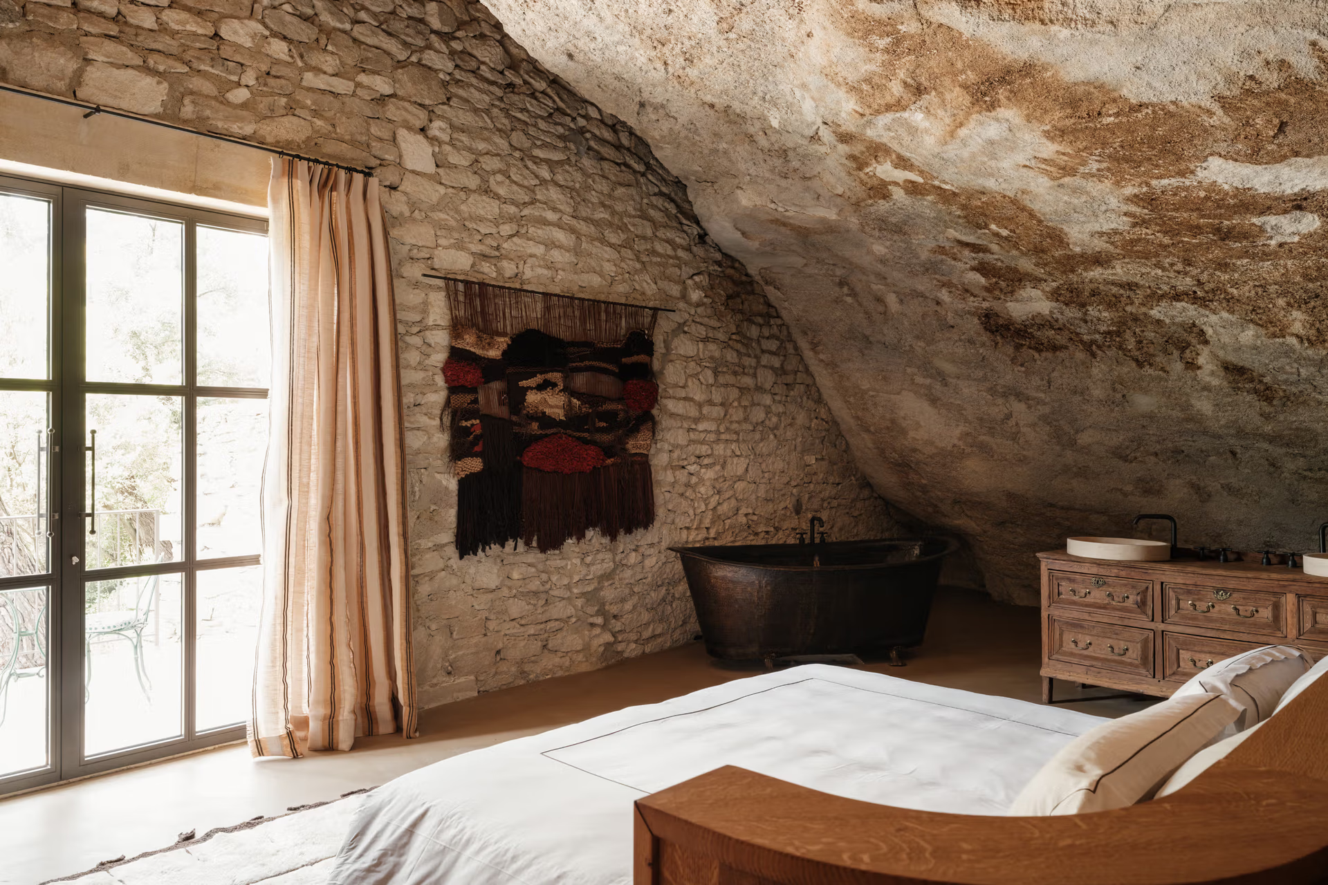 La Bergerie: rock-carved bedroom with curved limestone walls, black cast-iron soaking tub, wooden dresser, Berber textile art