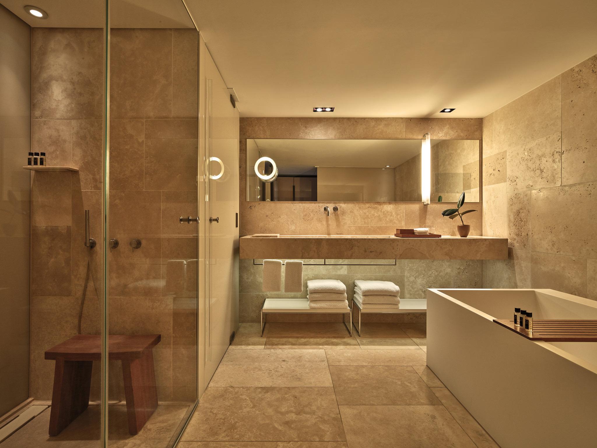 Mandarin Oriental Conservatorium, Amsterdam: stone-clad bathroom with a glass walk-in shower, a wooden stool, a white bathtub, and a lit mirror