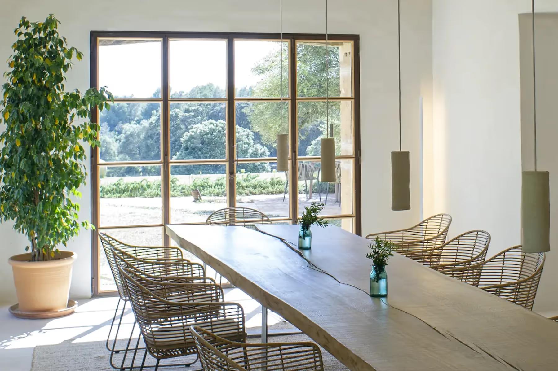 Contemporary dining room with long table, woven chairs, and full-height windows overlooking the estate