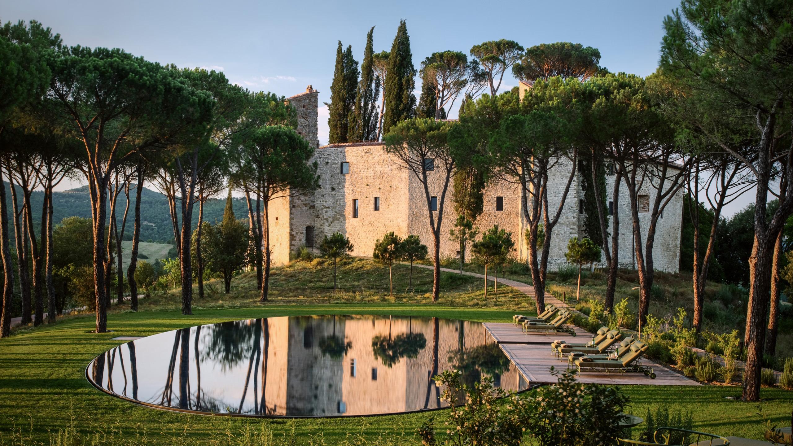 Castello di Reschio: the castle rising above pine trees, reflected in an oval pool cut into the lawn below