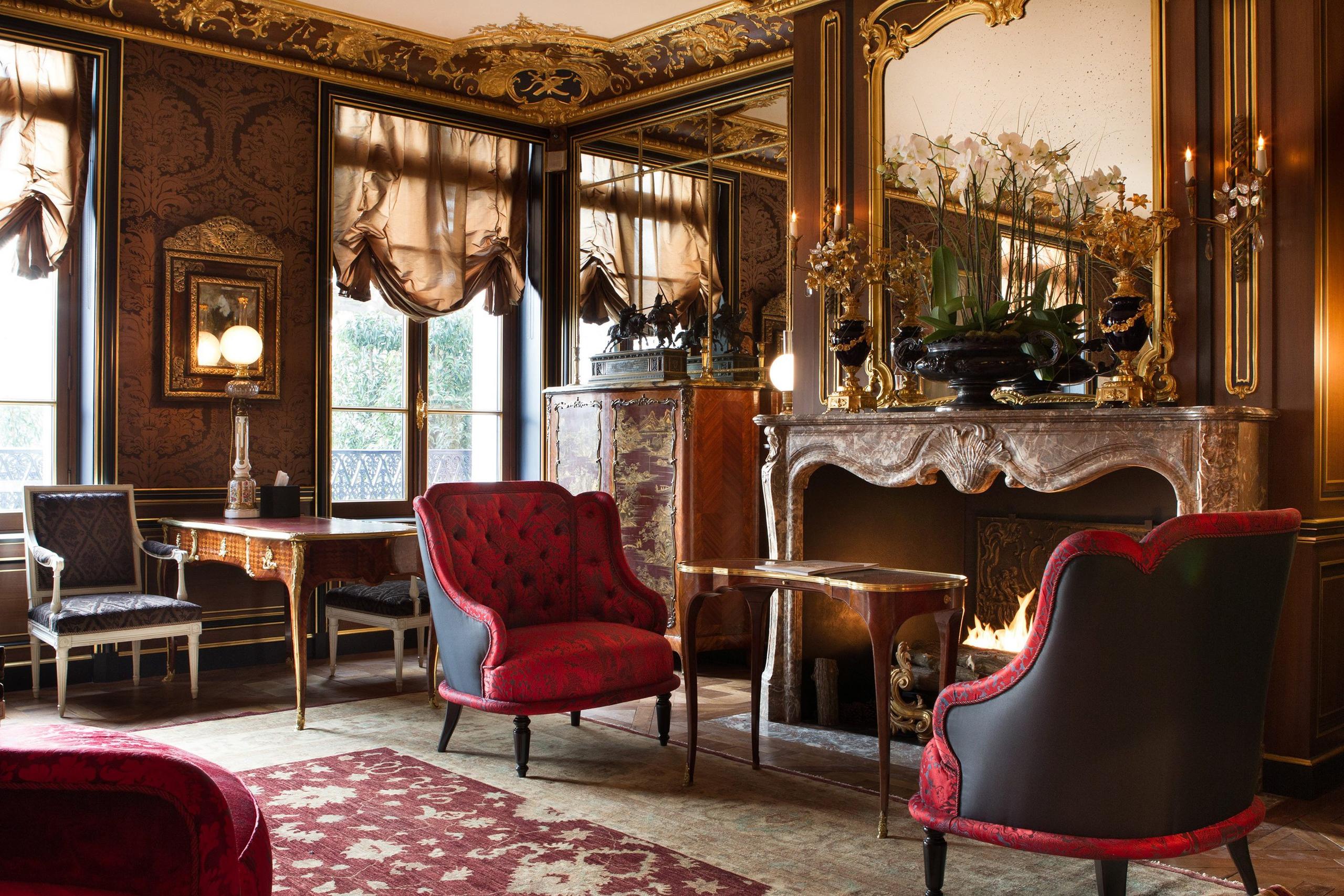 La Réserve Paris: a living room with cream and gold molding, burgundy furniture, and marble fireplace