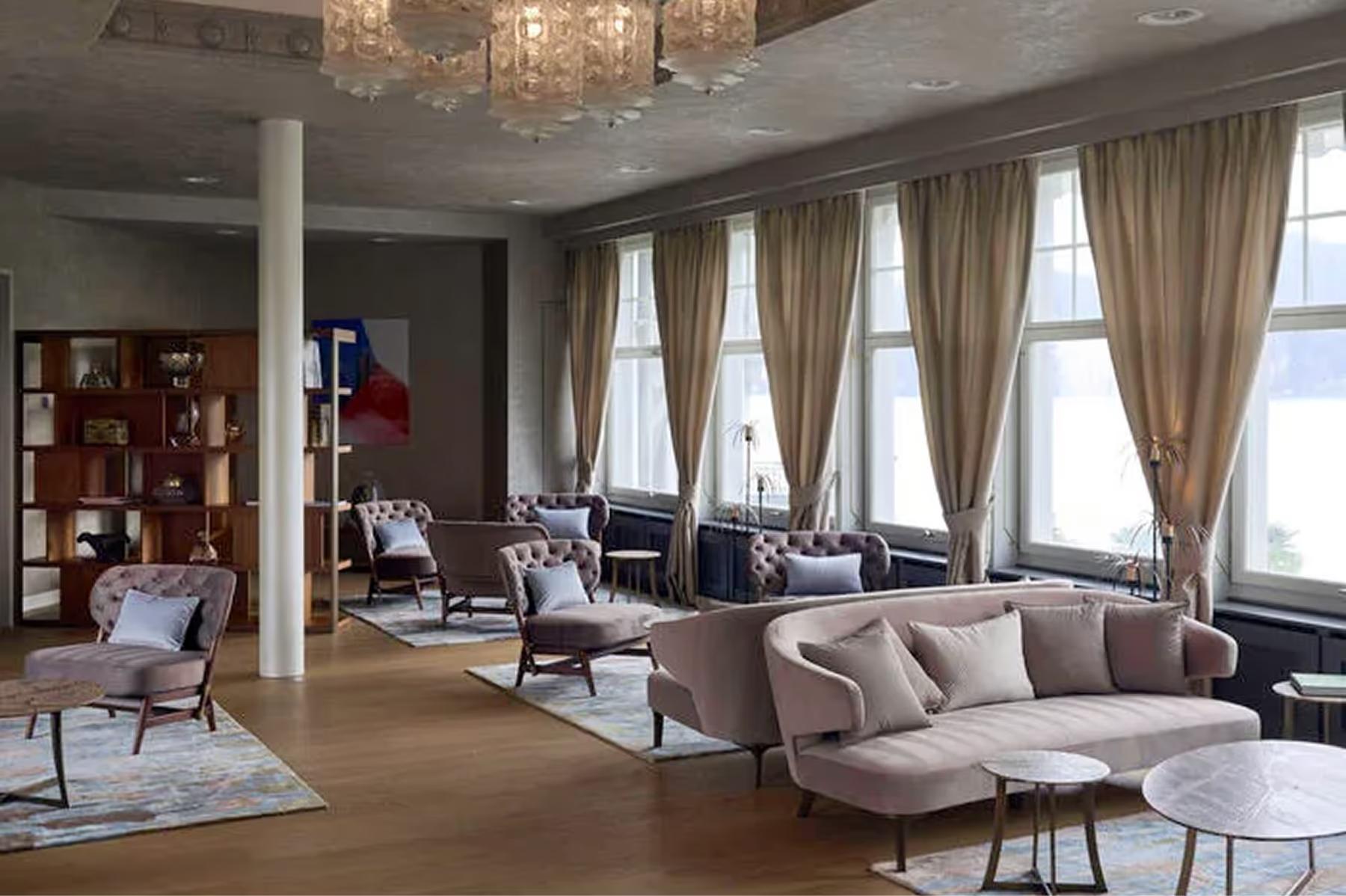 Chenot Palace Weggis: contemporary lounge with large windows, cream furnishings, wooden bookcase, and Alpine mountain view