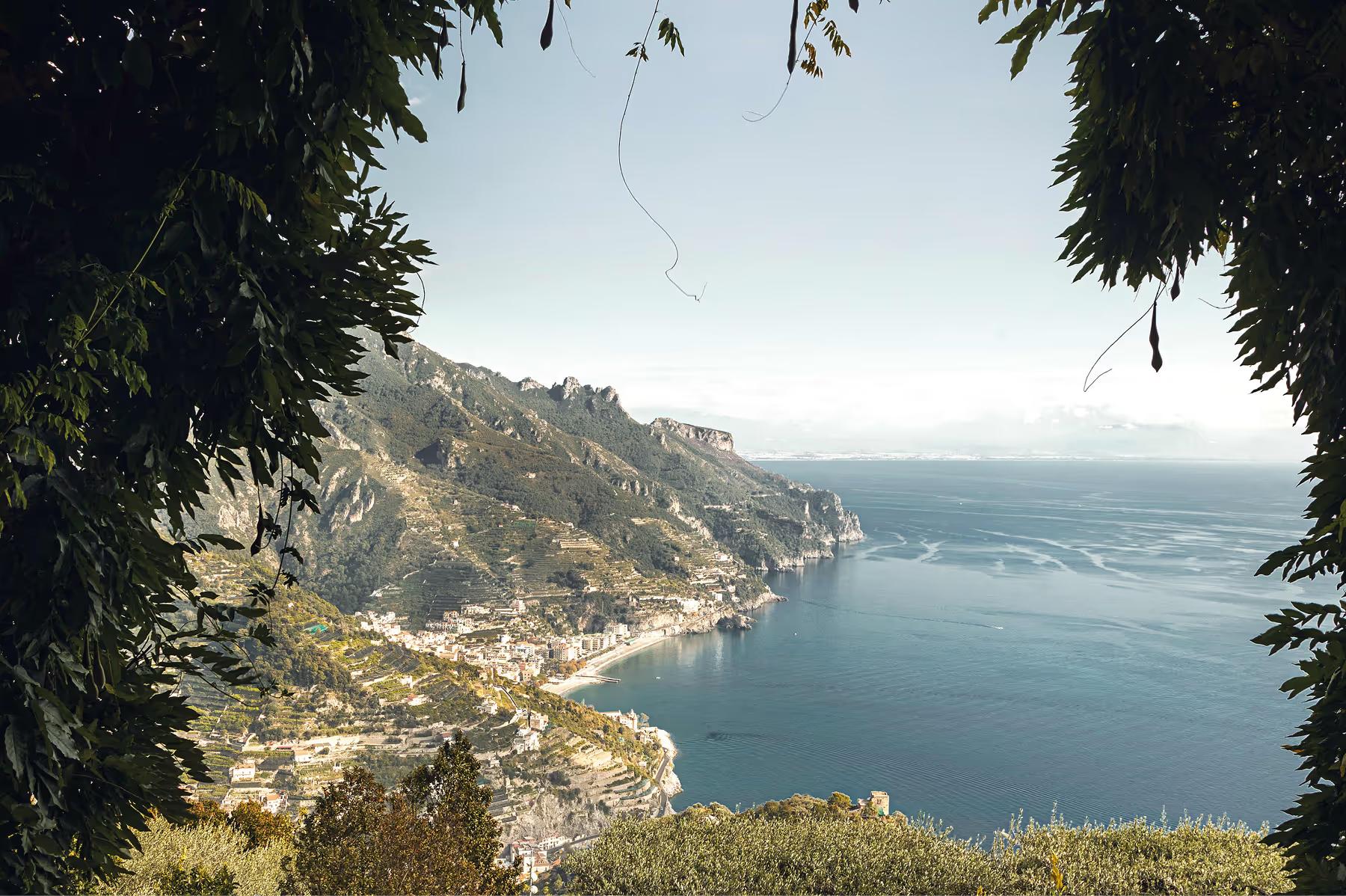 Caruso: coastal vista framed by hanging vines, turquoise sea meeting distant rocky hills under bright sky