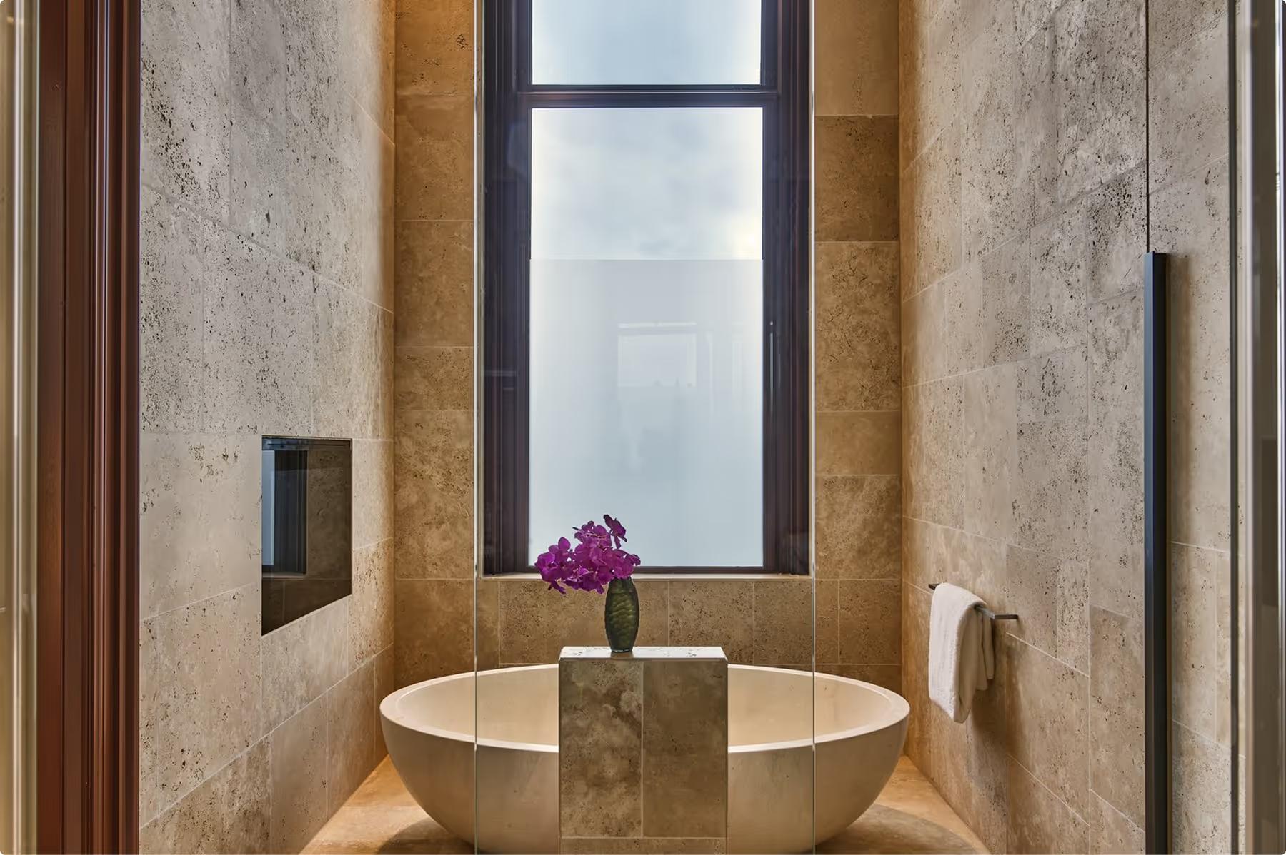 Mandarin Oriental Conservatorium, Amsterdam: stone-clad bathroom with an oval bathtub below a tall window and a vase of purple flowers