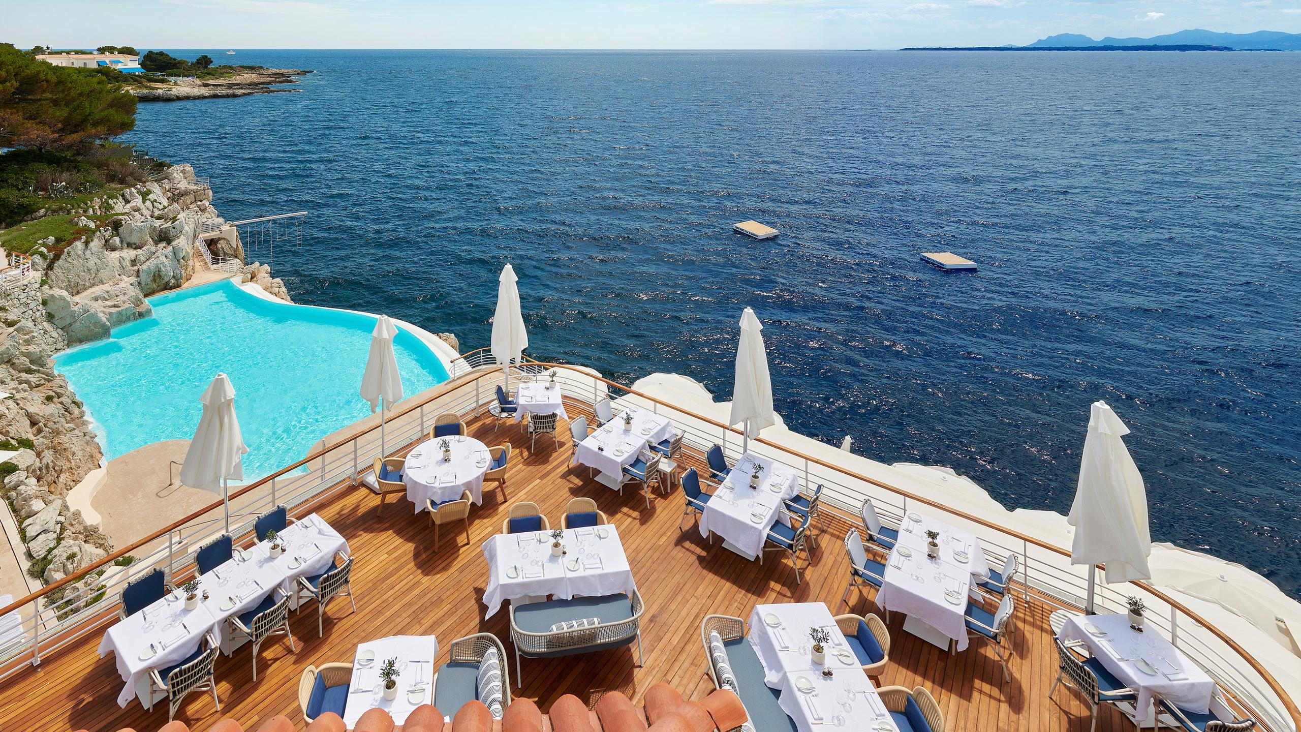 An open-air terrace restaurant with white umbrellas, sea views, and tables overlooking the Mediterranean