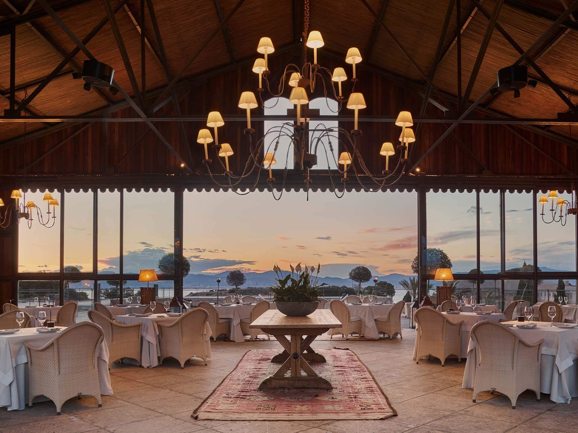 A vast dining pavilion with wooden beam trusses, brass lanterns, cream chairs, and panoramic sea views
