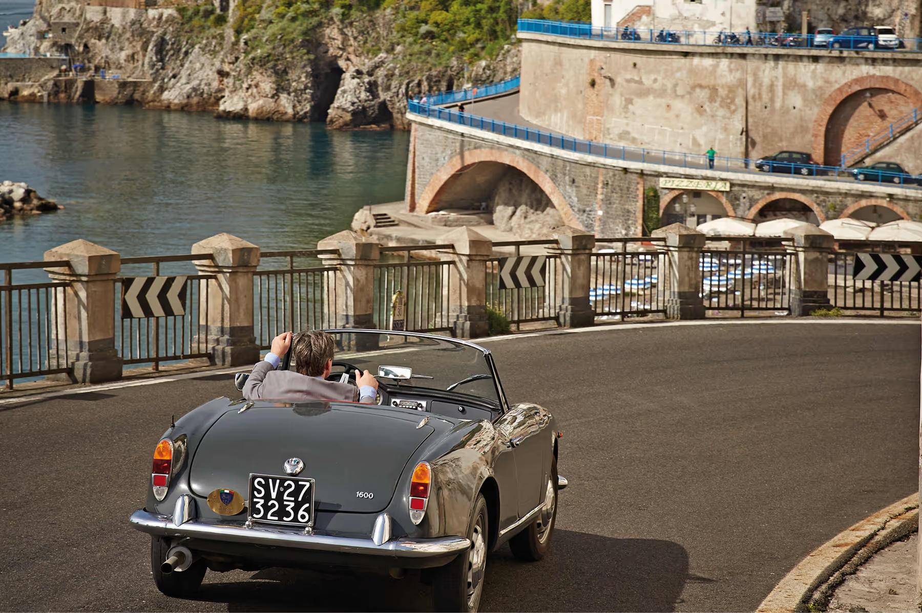 Caruso: vintage convertible car parked on coastal bridge with waterfront buildings and boats