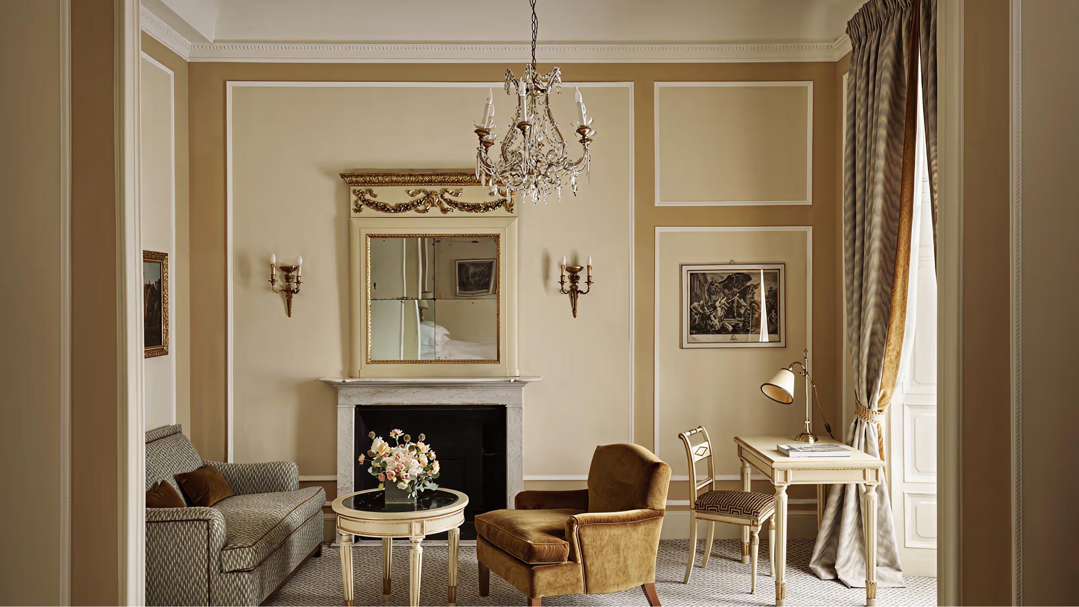 A formal sitting room with cream walls, fireplace, crystal chandelier, and gilded accents