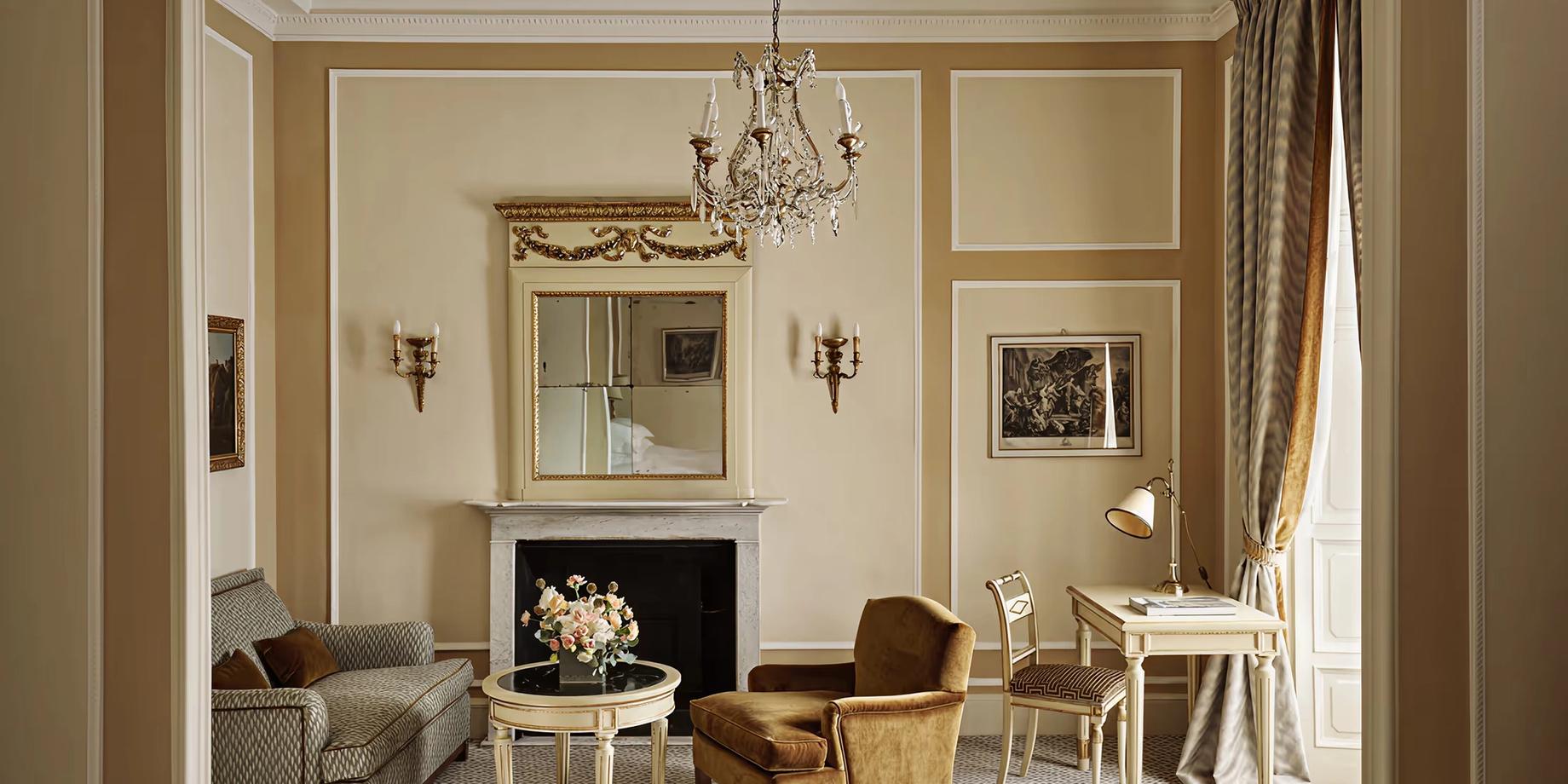 A formal sitting room with cream walls, fireplace, crystal chandelier, and gilded accents