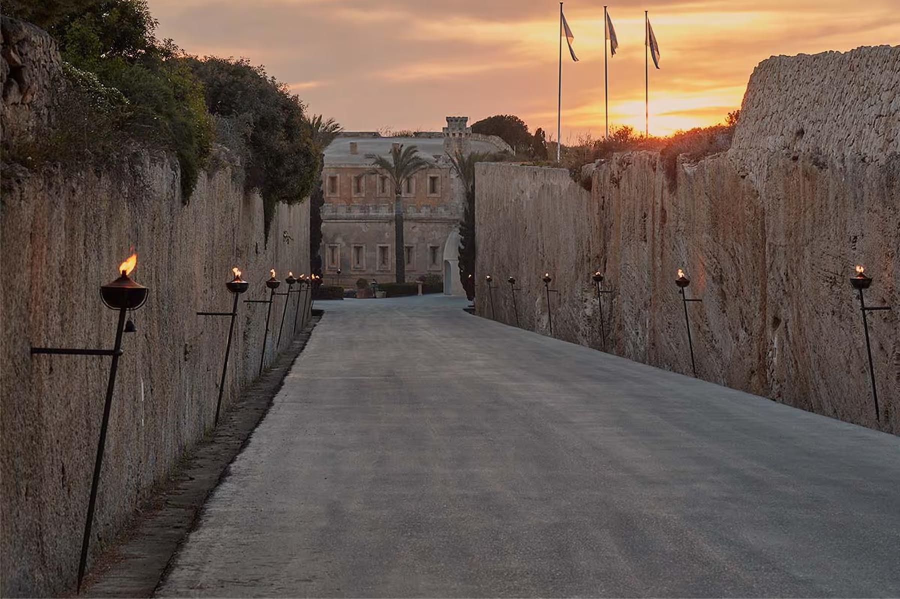 A stone causeway bordered by fortress walls at dusk, leading toward the Mediterranean