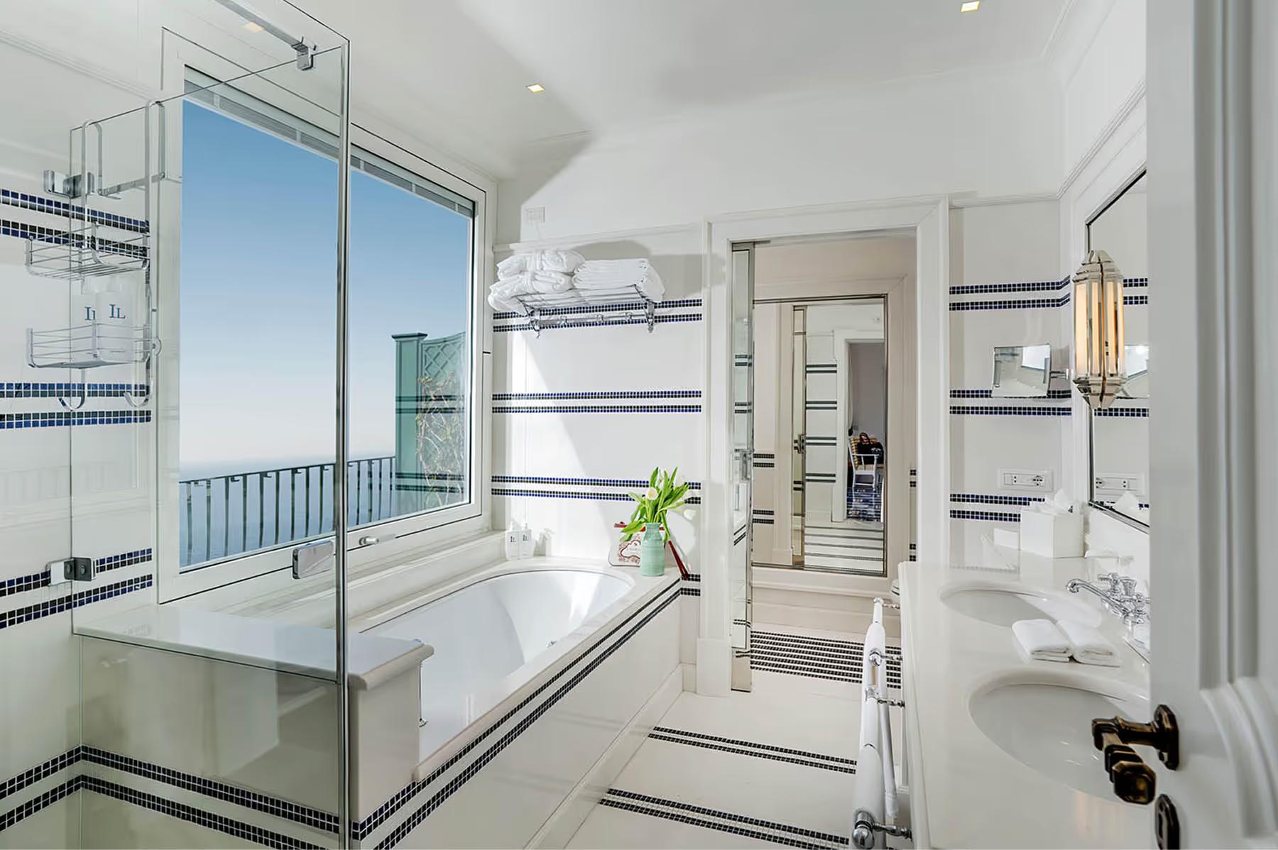 Il San Pietro di Positano: white bathroom with glass shower enclosure, spa tub, marble accents, and view of sea beyond