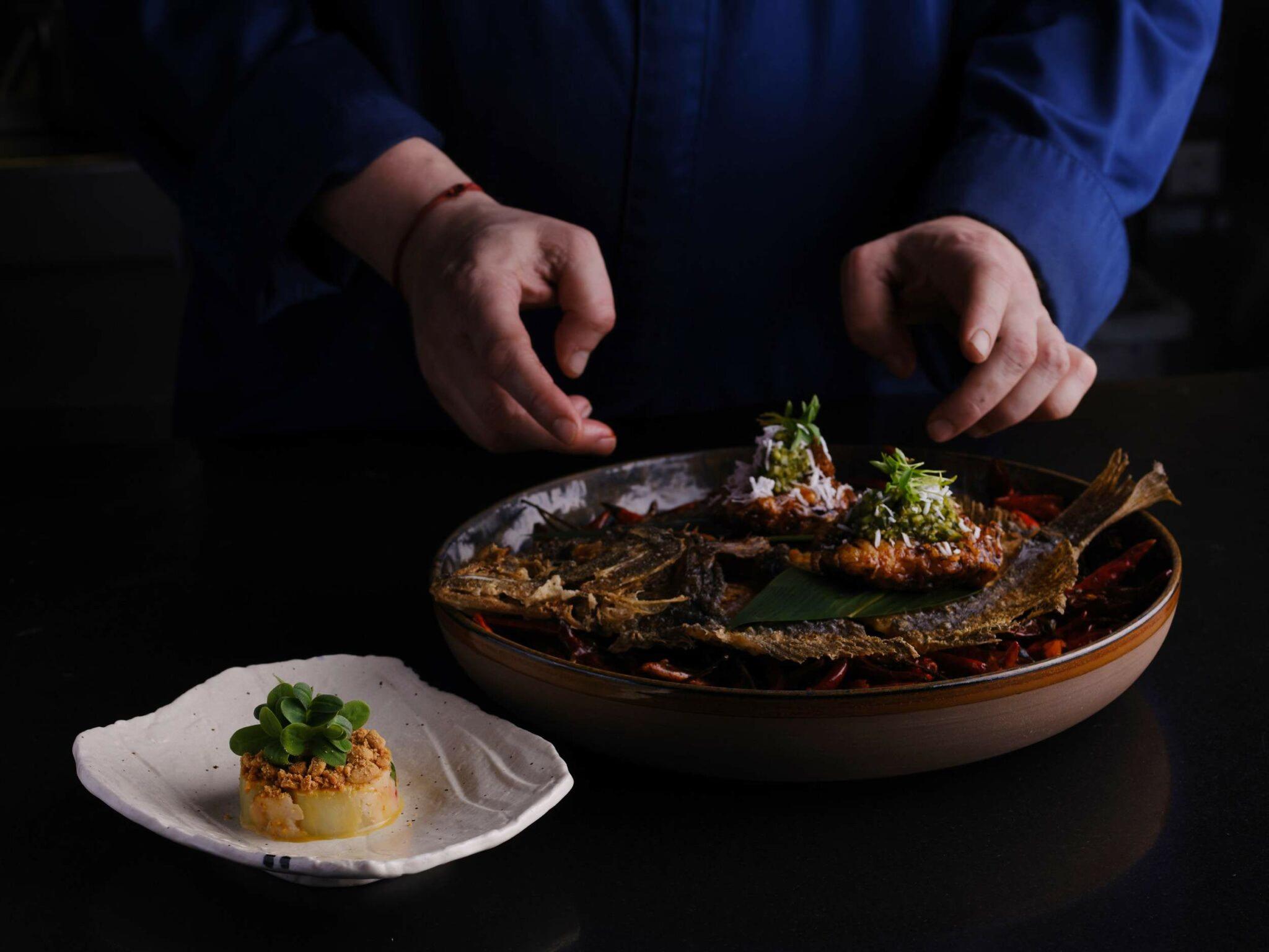 Mandarin Oriental Conservatorium, Amsterdam: Pan-Asian seafood dish plated on slate with wooden bowl and garnish, hands presenting the plate