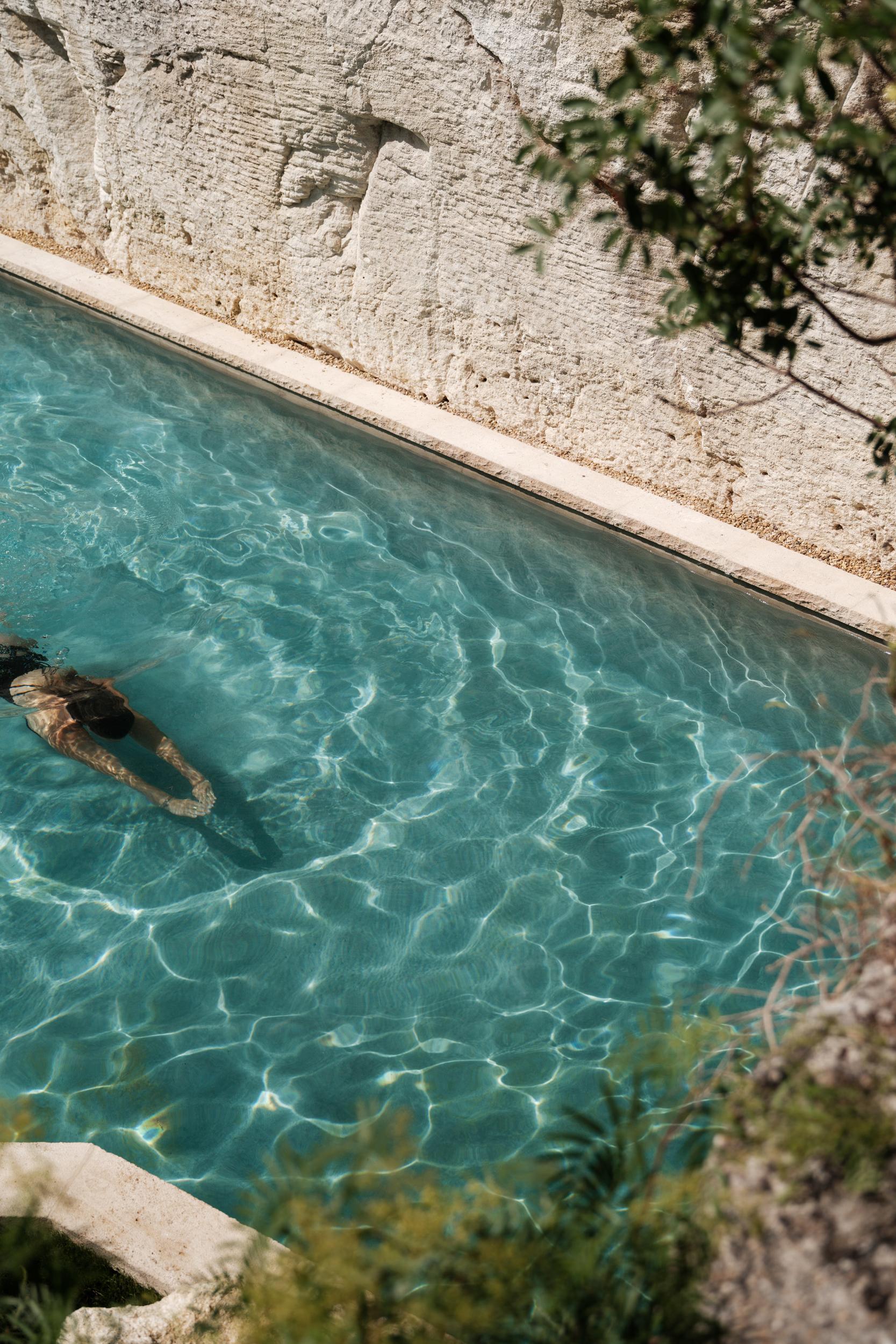 La Bergerie: swimmer in crystal-clear turquoise pool beside textured limestone wall, bright water refraction patterns