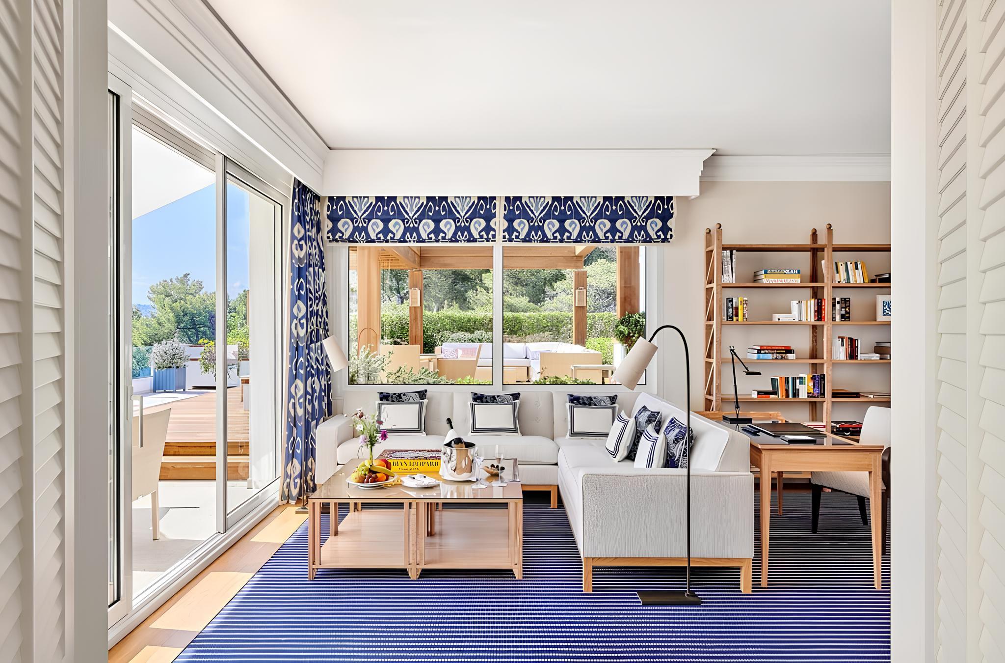 A sitting room with blue carpet, linen furniture, arched windows opening to sunlit grounds