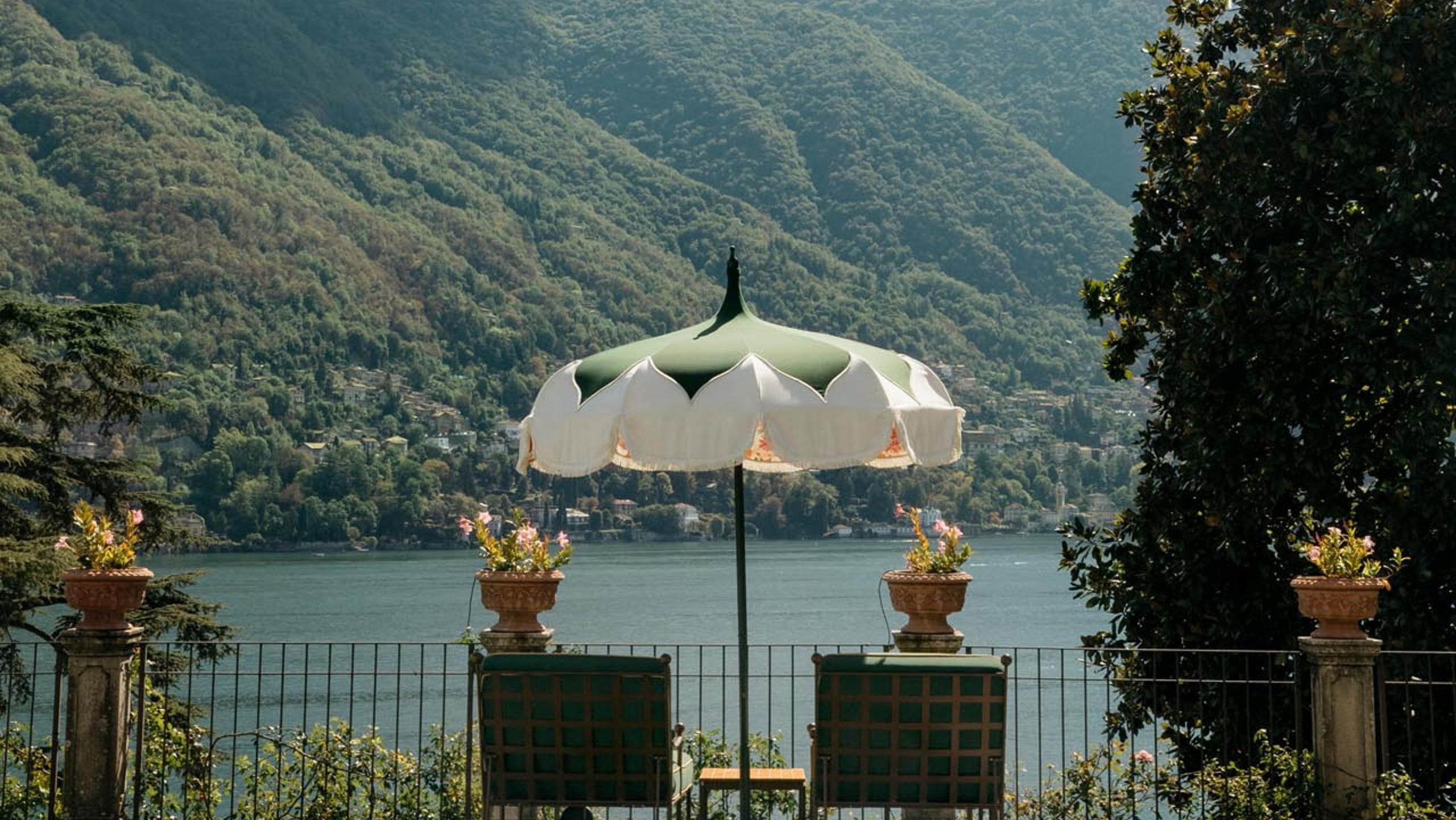 A cream umbrella and green chairs on a terrace overlooking Lake Como at golden hour
