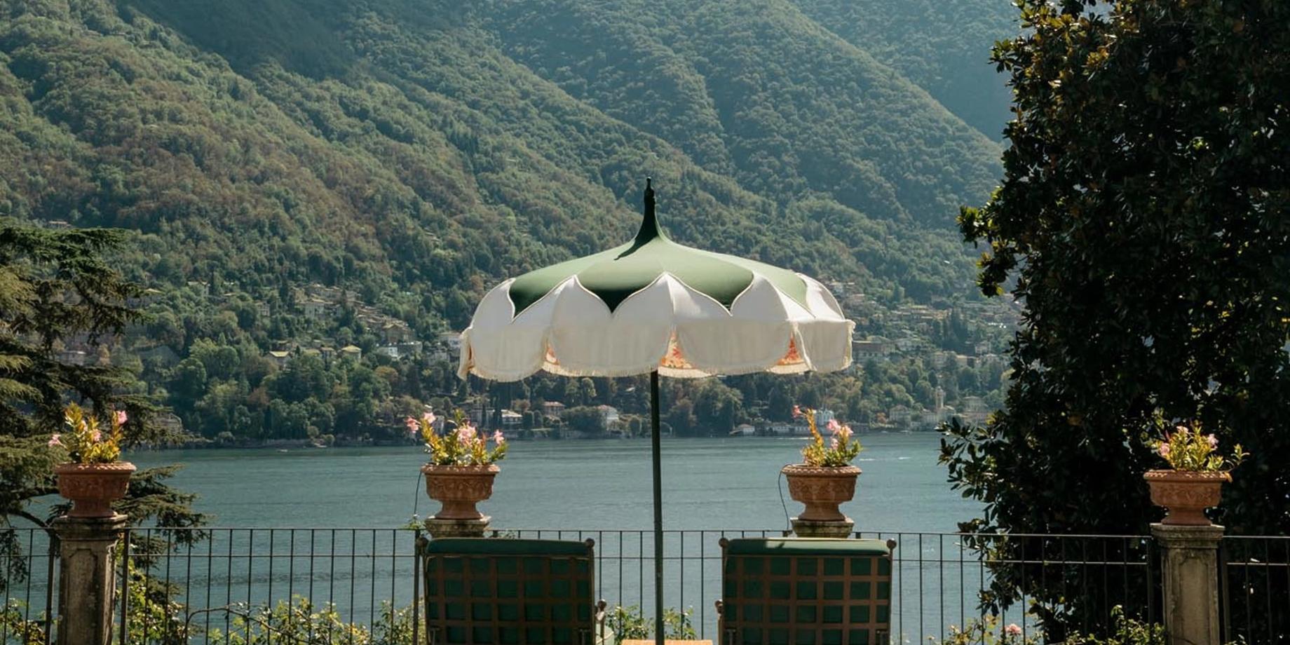 A cream umbrella and green chairs on a terrace overlooking Lake Como at golden hour