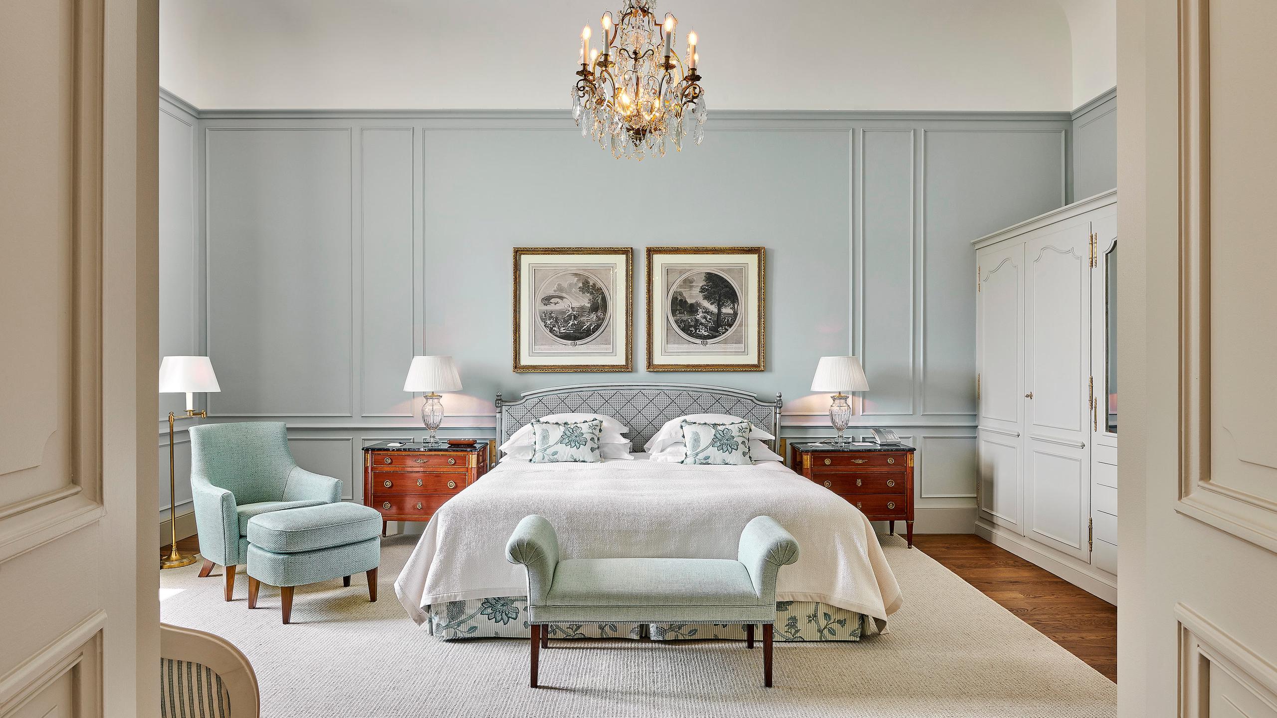 A guest bedroom with soft green walls, a white bed, matching armchairs, and a crystal chandelier