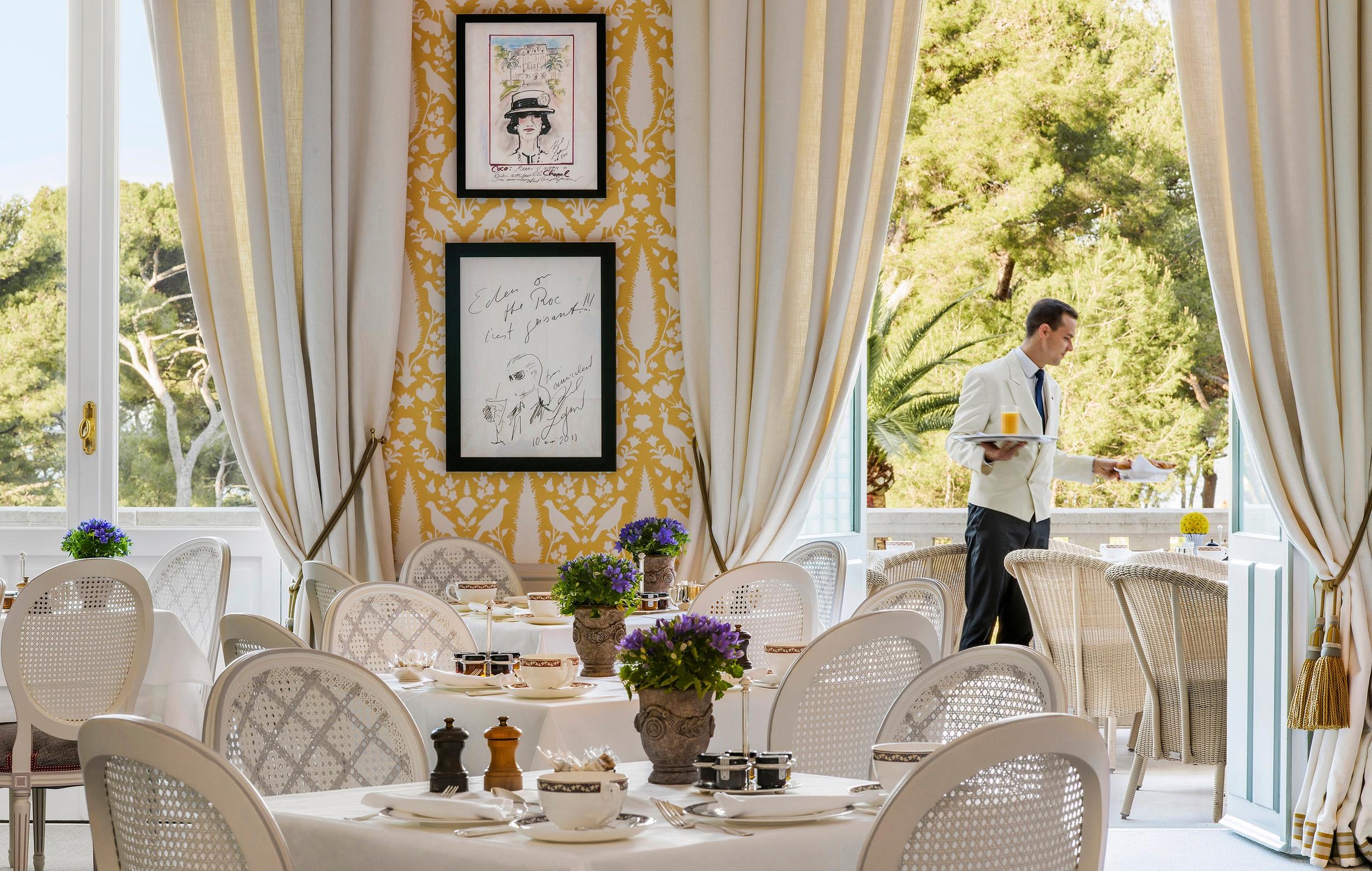 An elegant dining room with cream drapery, framed artwork, white tablecloths, and garden views
