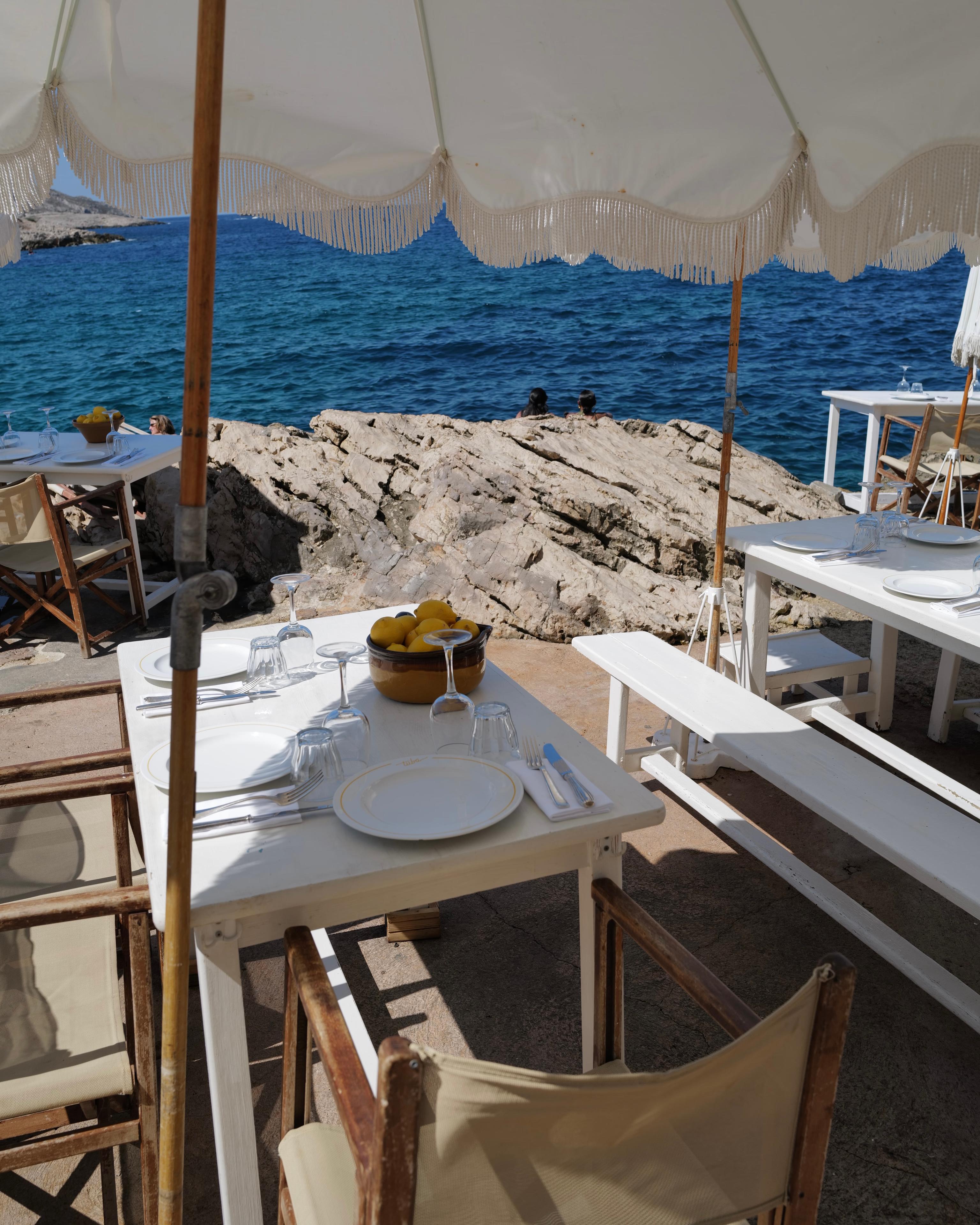 A set table under a fringed parasol at Tuba Club, the rocks and open sea beyond
