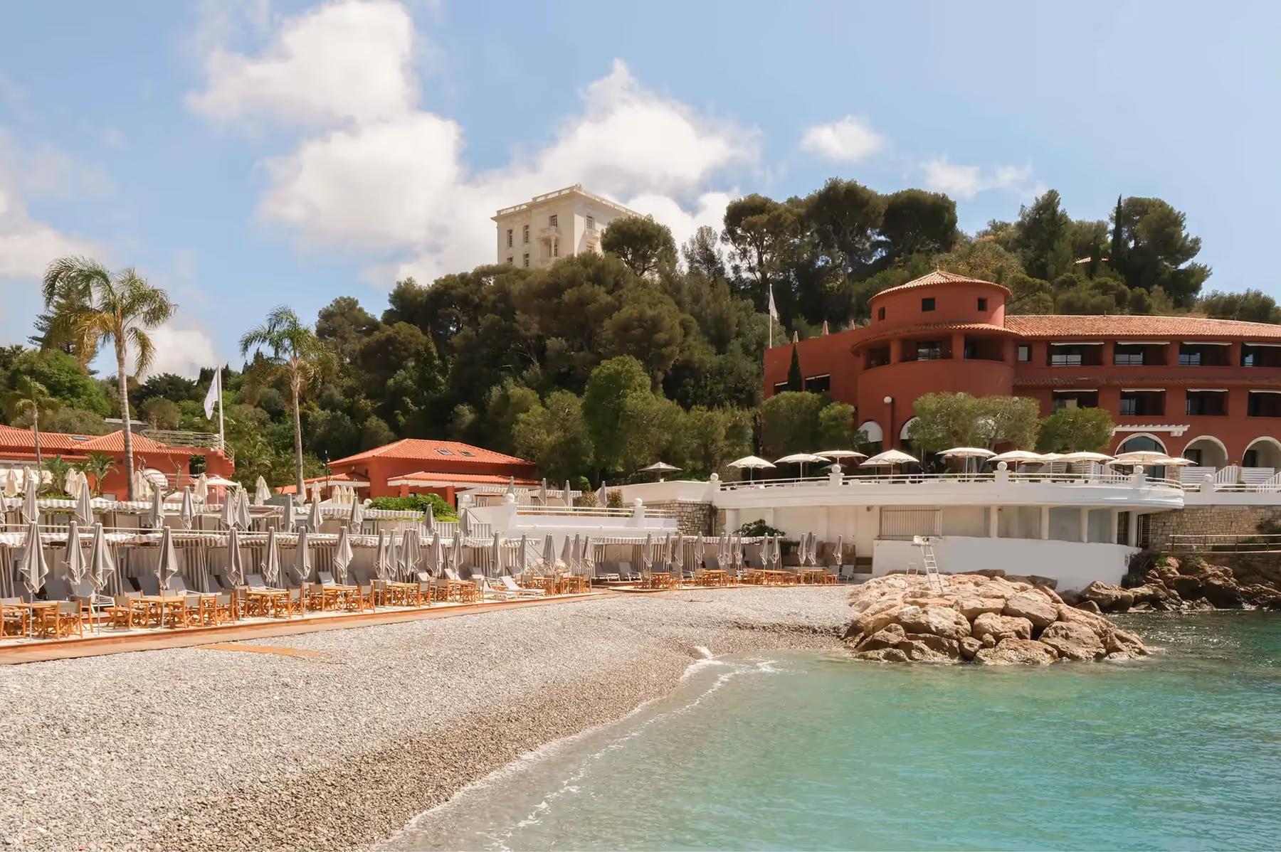 Pebble beach with white loungers and umbrellas, turquoise Mediterranean water, terracotta hotel buildings backed by pine trees