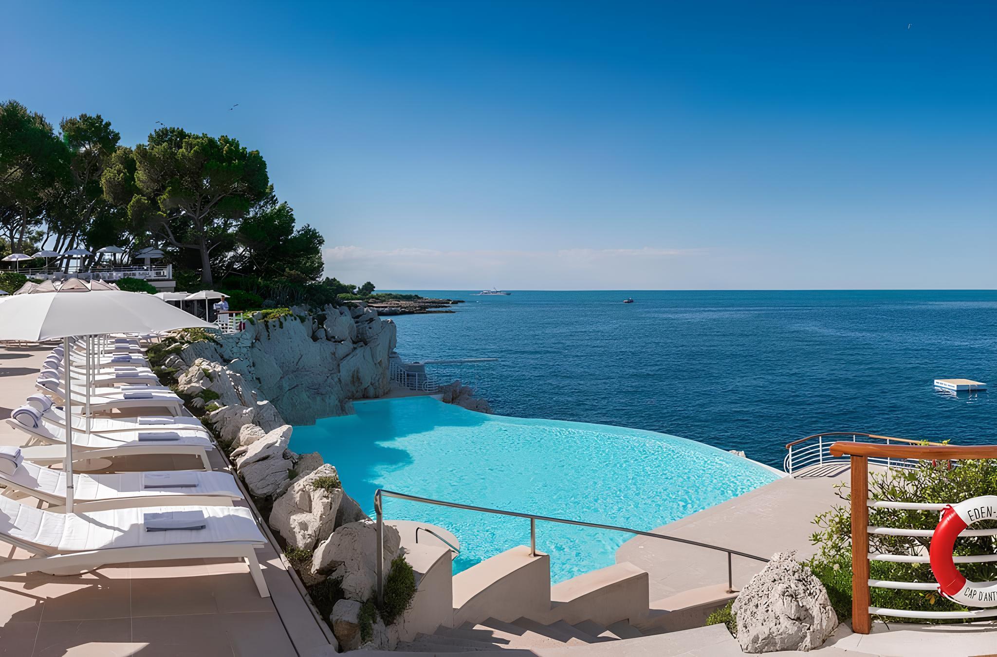A turquoise infinity pool above the sea, with loungers, bright sun umbrellas, and rocky coastline