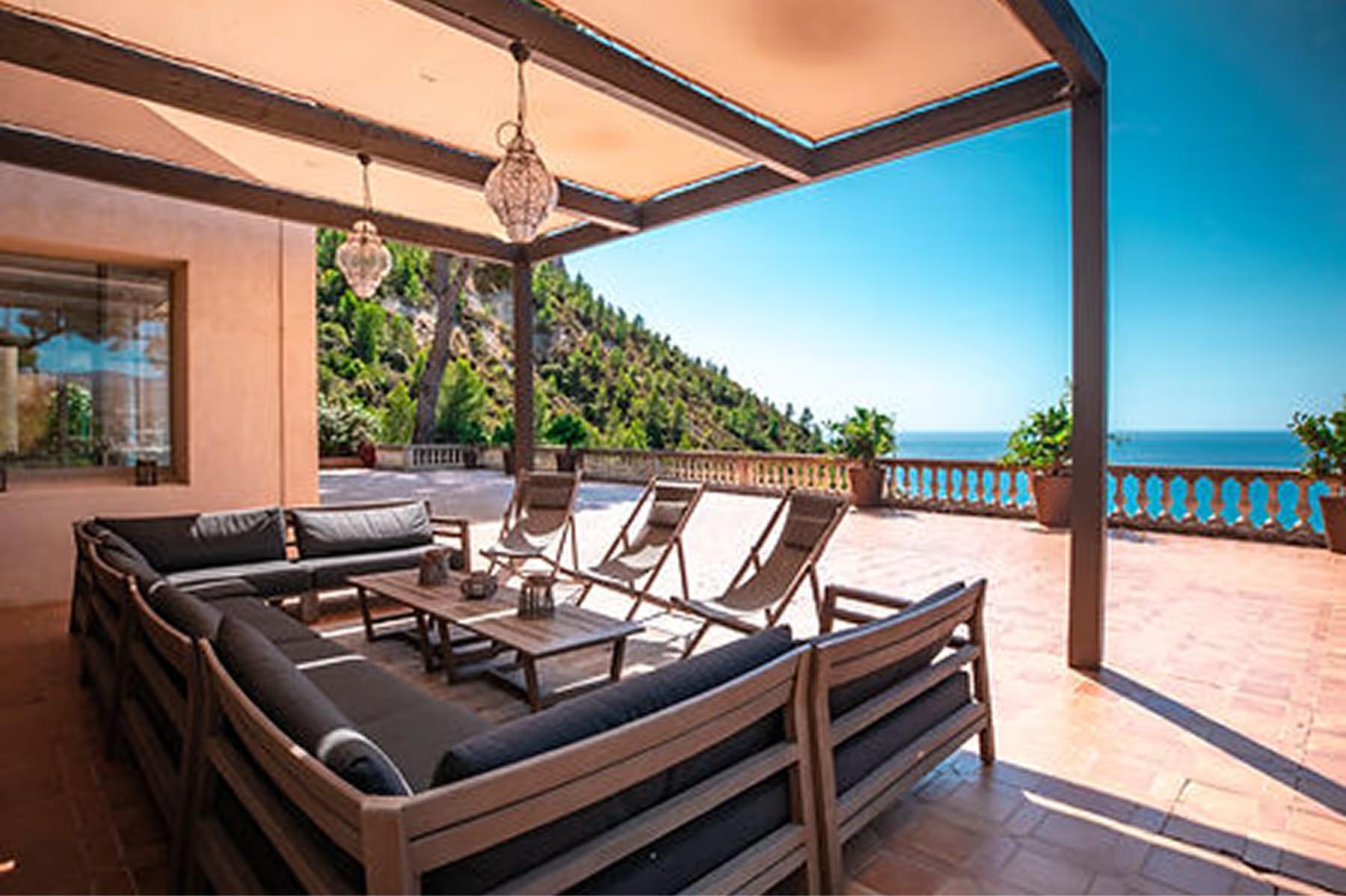 Covered terrace with dark wooden furniture and cushions overlooking turquoise sea and pines