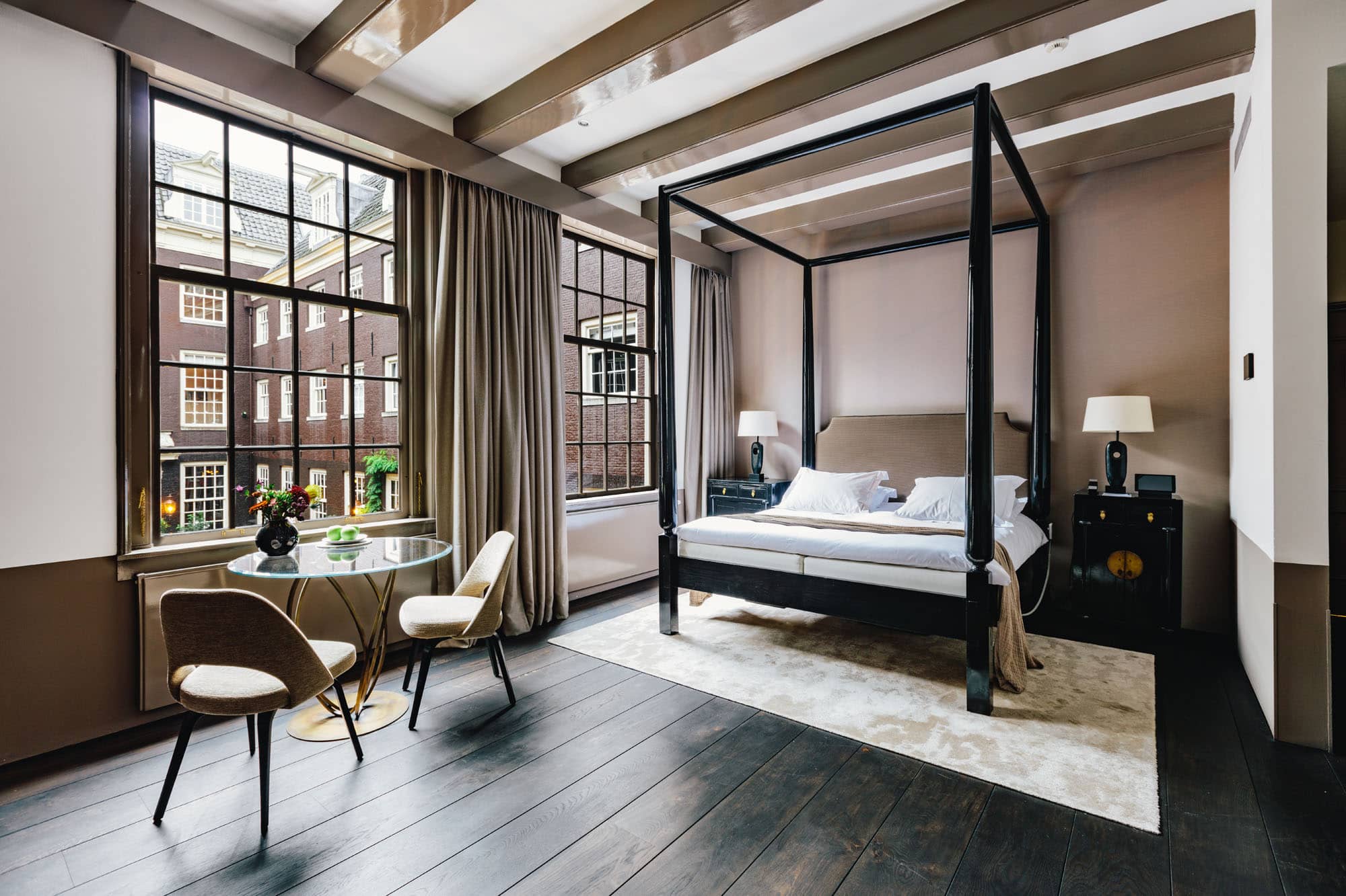 A four-poster bed under the beams at The Dylan, with a small glass table at the windows onto the canal houses
