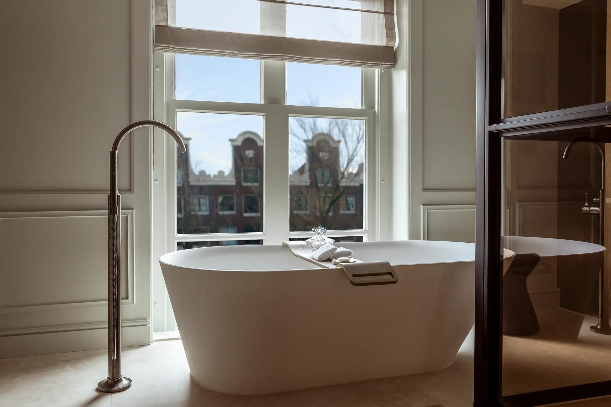 Rosewood Amsterdam: a Canal View Executive Suite bathroom with a freestanding tub at a window over gabled houses
