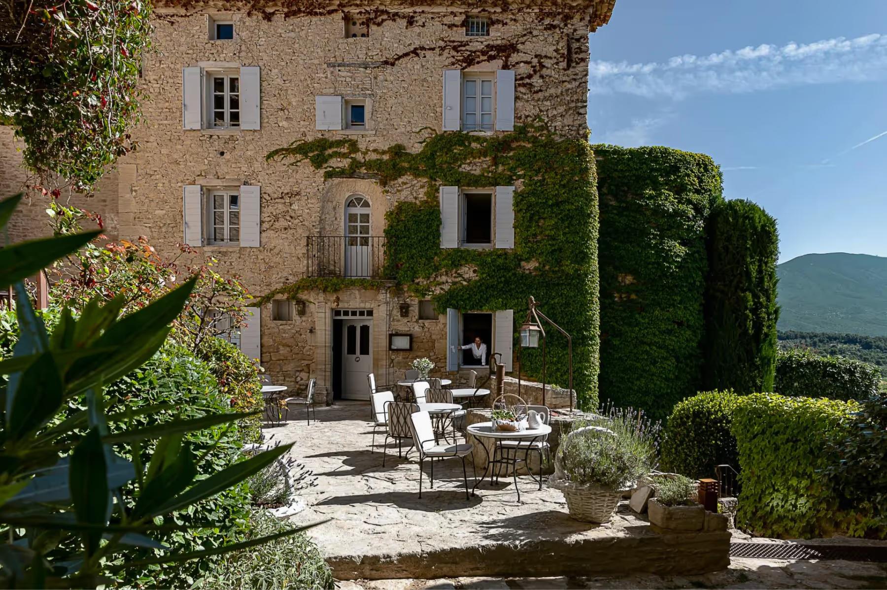 Stone building with climbing ivy, white shutters, dining terrace, potted plants, cypress hedges, and mountain vista