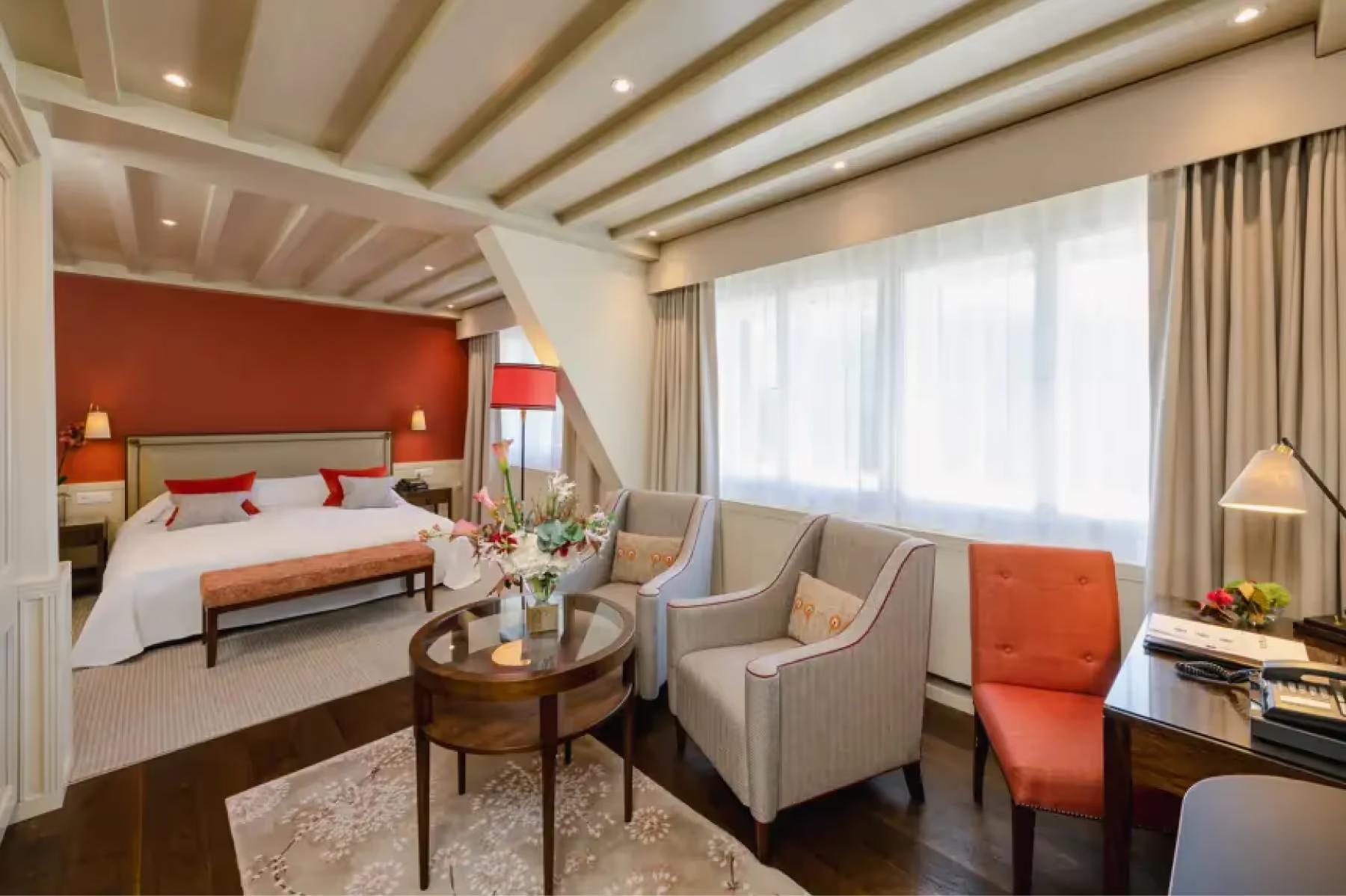 Suvretta House: a bedroom with red headboard, lounge seating, wood beams, and bay windows with mountain views