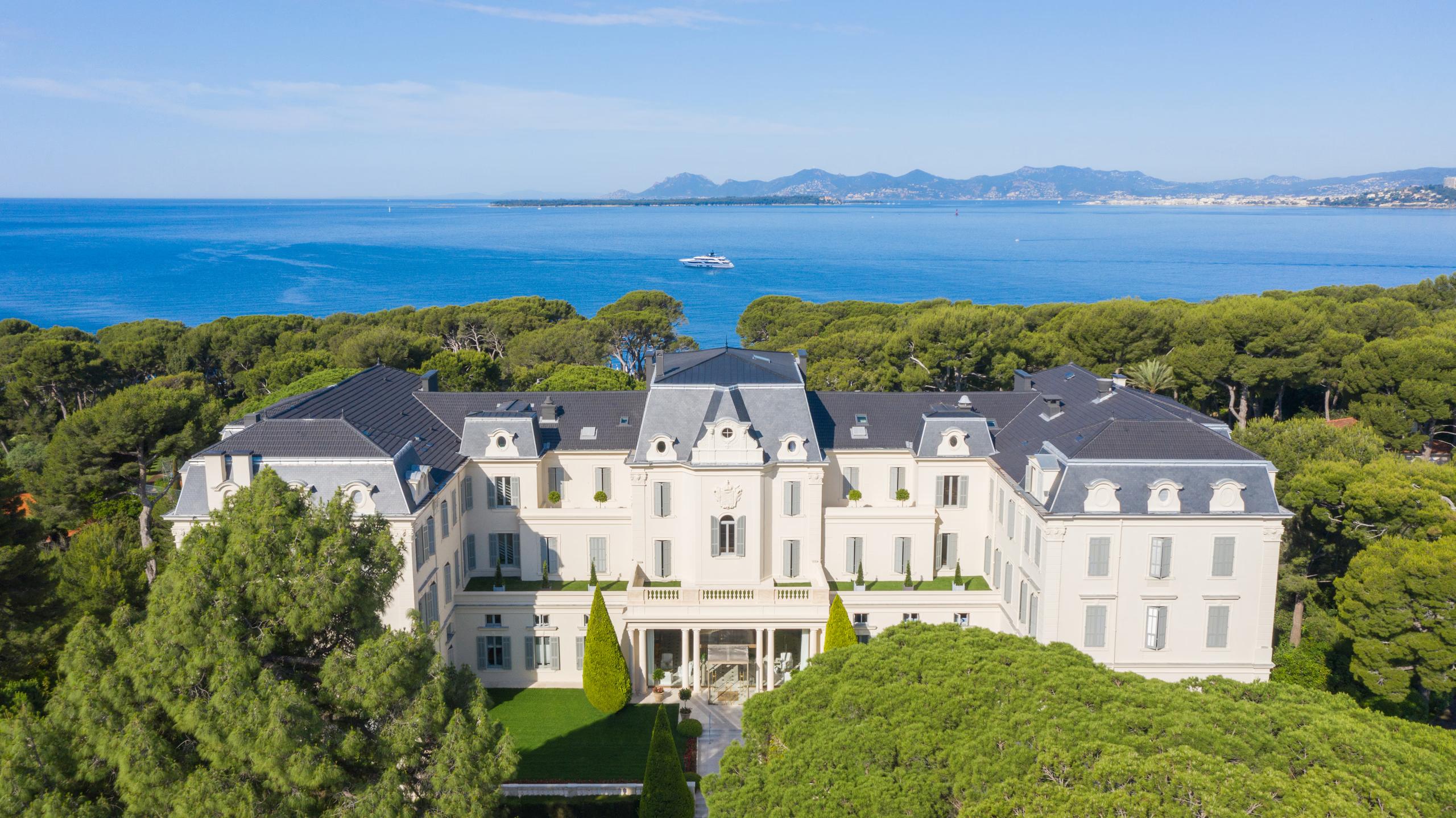 An aerial view of a cream-colored mansion with mansard roofs overlooking the Mediterranean and distant coastline