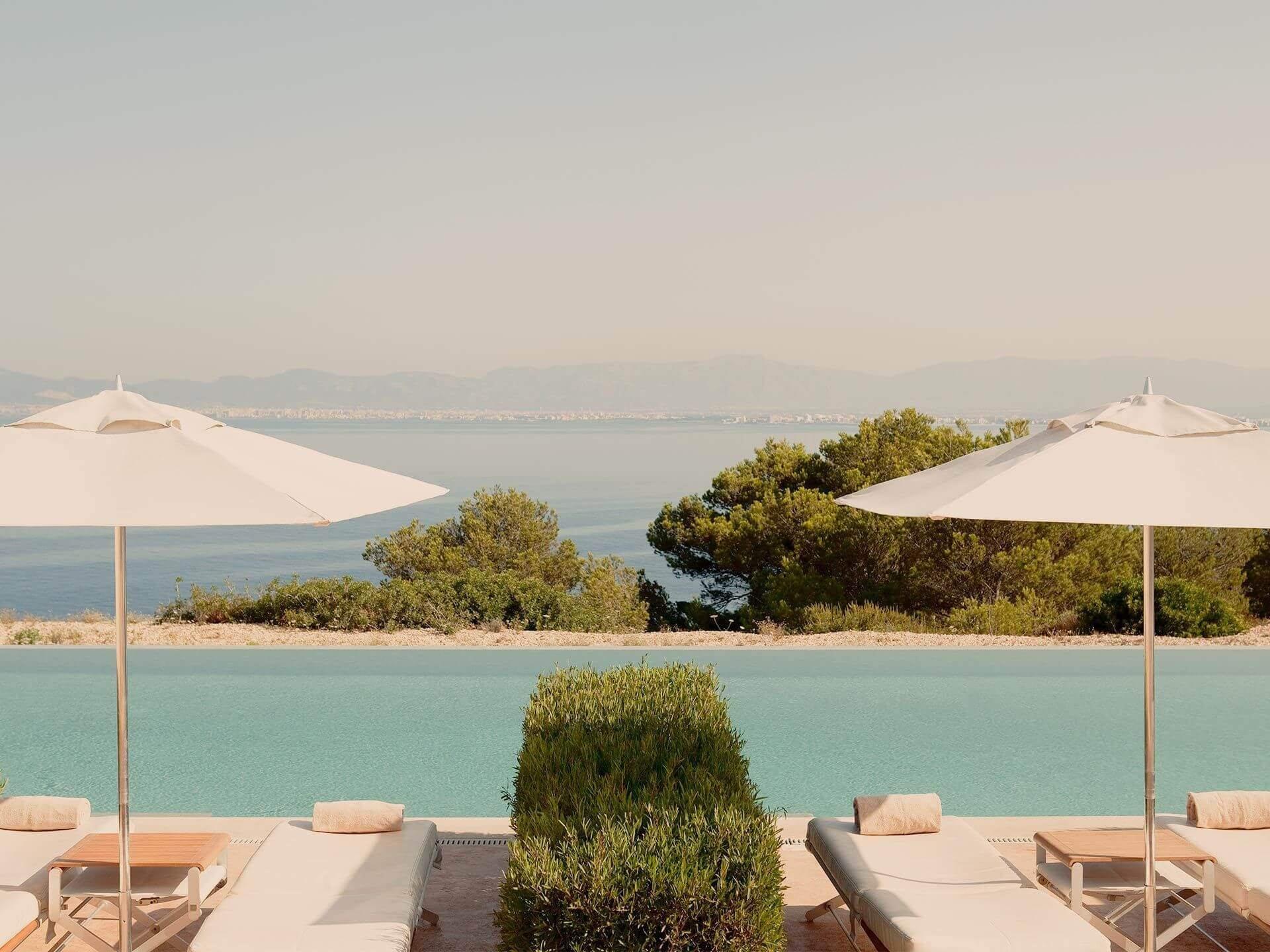 A saltwater pool edged with cream loungers and white parasols, overlooking calm turquoise bay