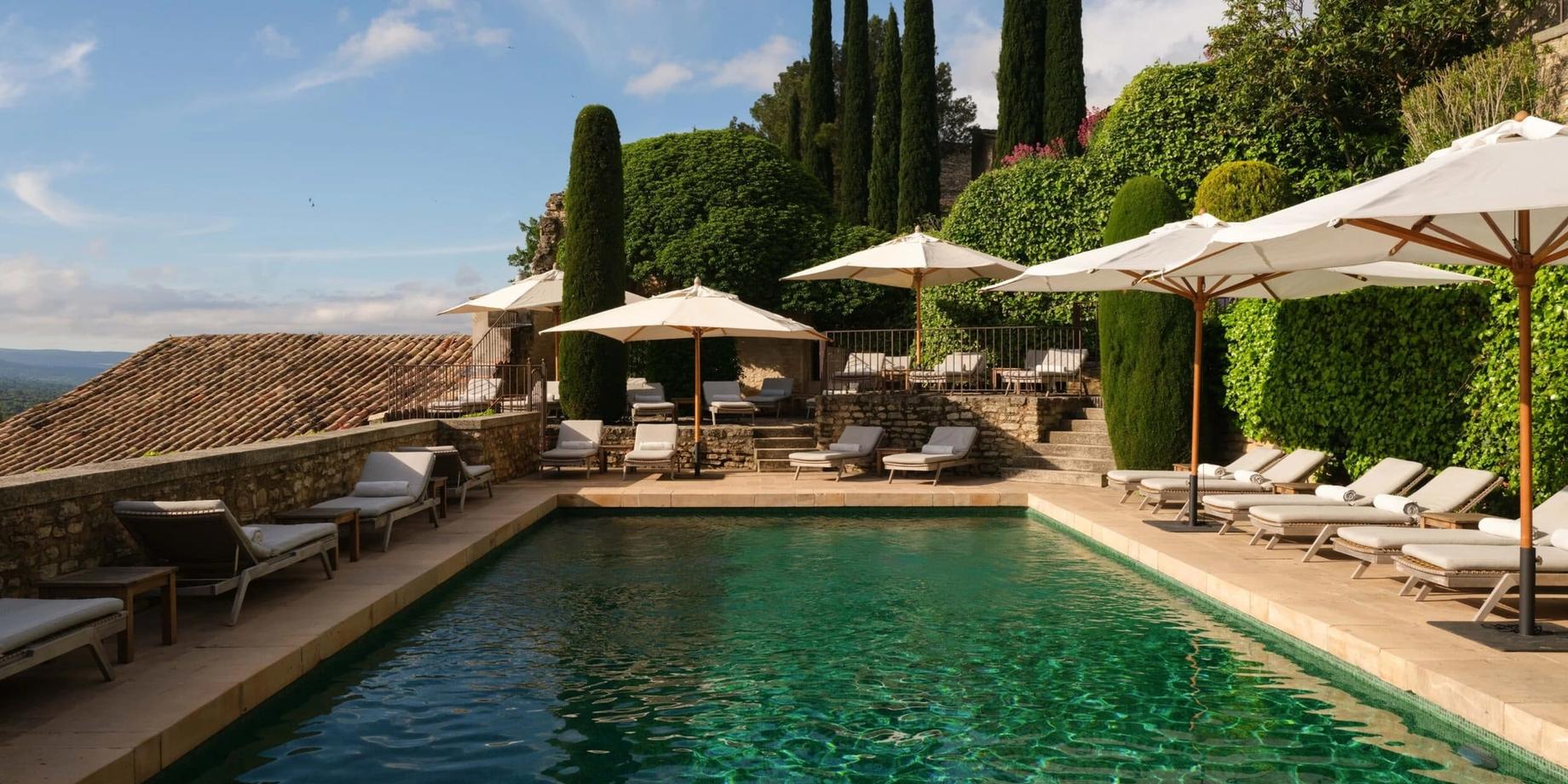 A swimming pool overlooking rolling vineyards and mountains under a blue sky with cypress trees and lounging areas