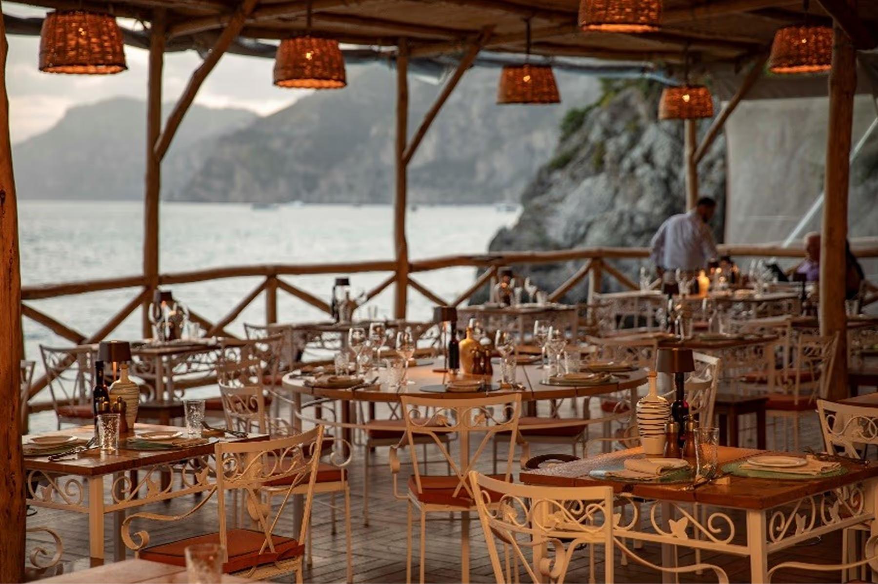 Il San Pietro di Positano: covered restaurant interior with wooden chairs, round tables, white linens, and Mediterranean coastline backdrop