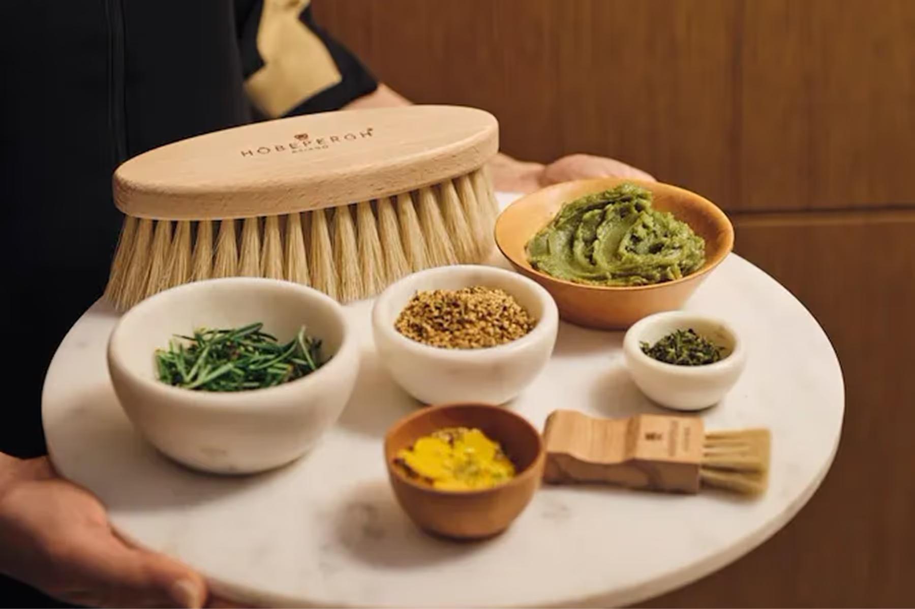 Spa treatment items in white bowls on a table with natural ingredients and a wooden brush