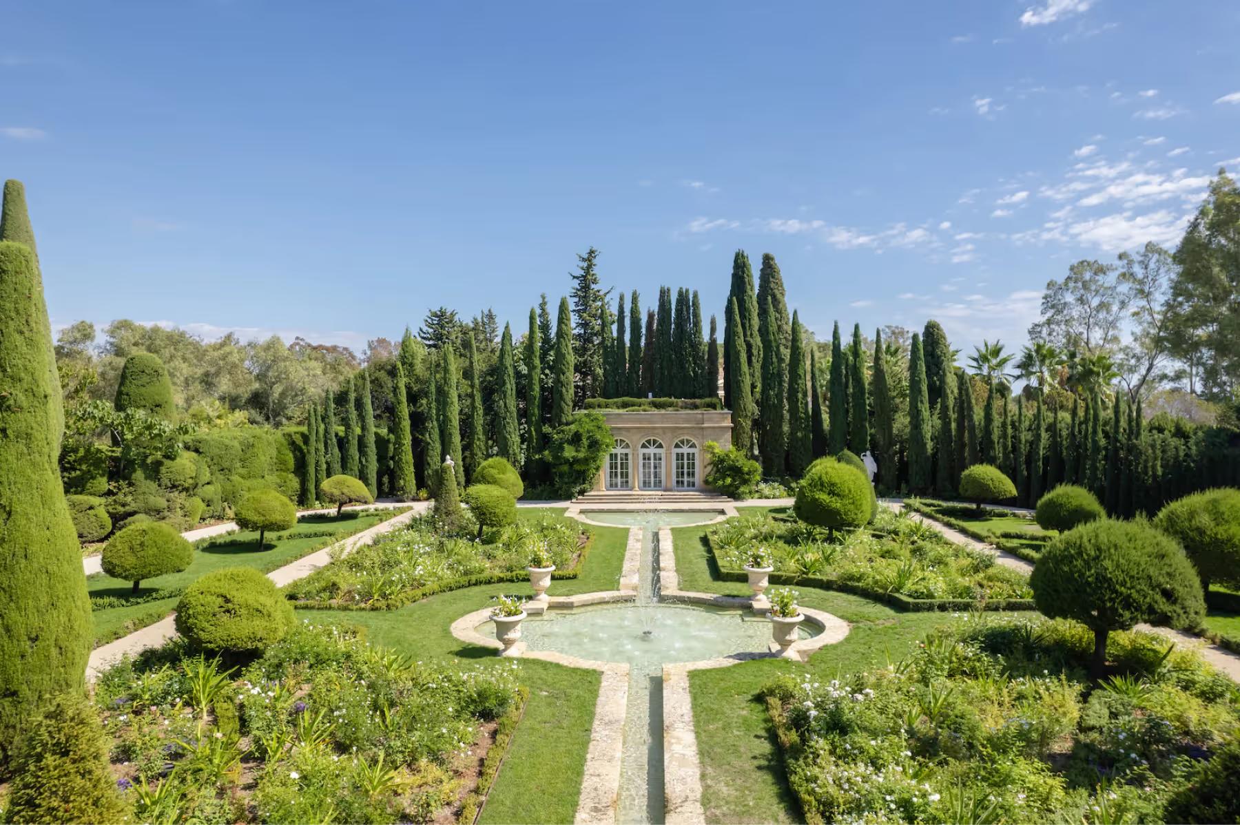 Formal gardens viewed from above, with stone pathways, cypress trees, and château roofline